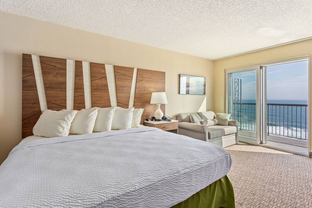 Een hotelkamer met een groot bed, een sofa, een nachtkastje en uitzicht op de oceaan vanaf een balkon.