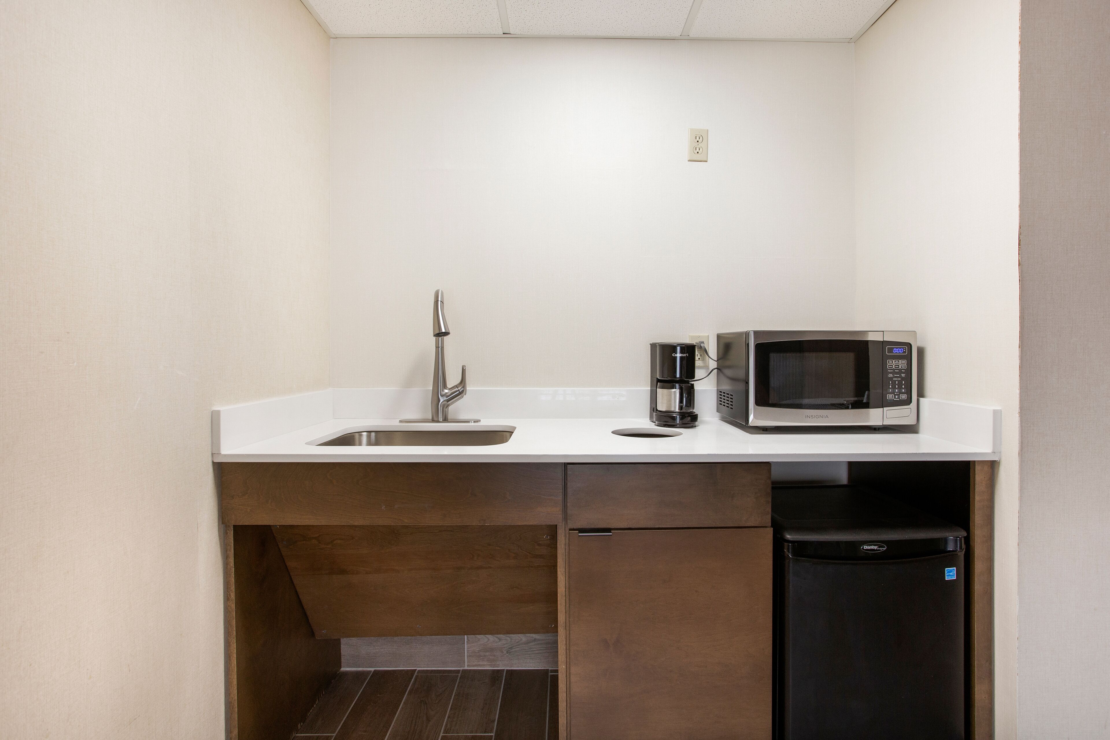 A compact kitchenette with a white countertop, stainless steel sink, a coffee maker, a microwave, and a black dishwasher.