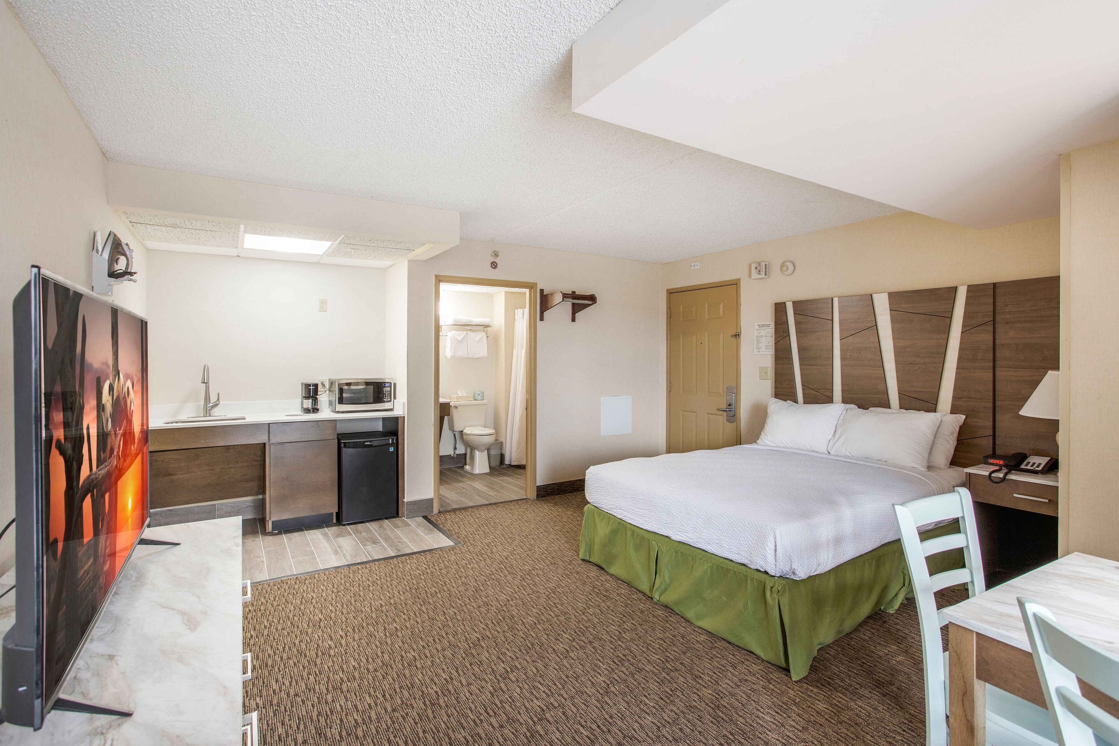 Habitación de hotel con cama, escritorio, kitchenette y baño visible a través de una puerta abierta.