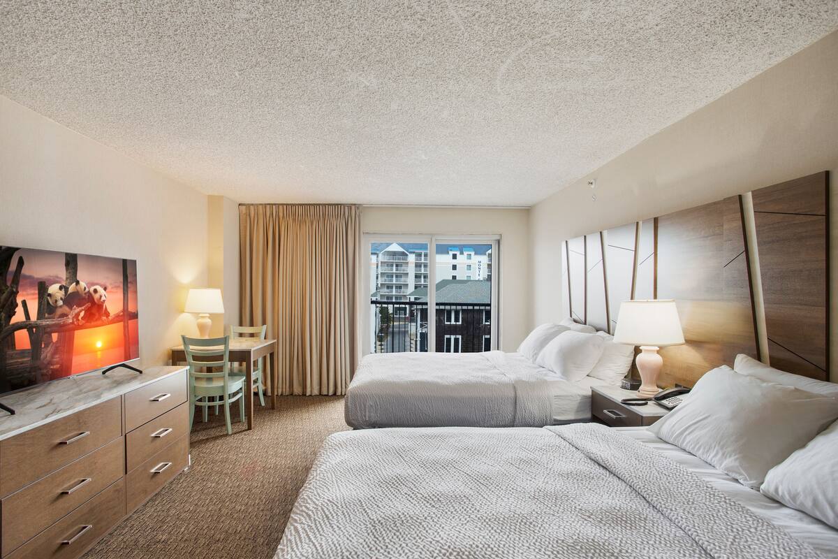 Hotelkamer met twee bedden, een televisie, een bureau met stoel en een raam met uitzicht op gebouwen.