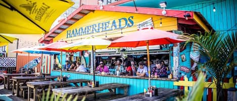 Een terras buiten met houten tafels en banken, een kleurrijk gebouw met het bord 'HAMMERHEADS', en parasols voor de schaduw.