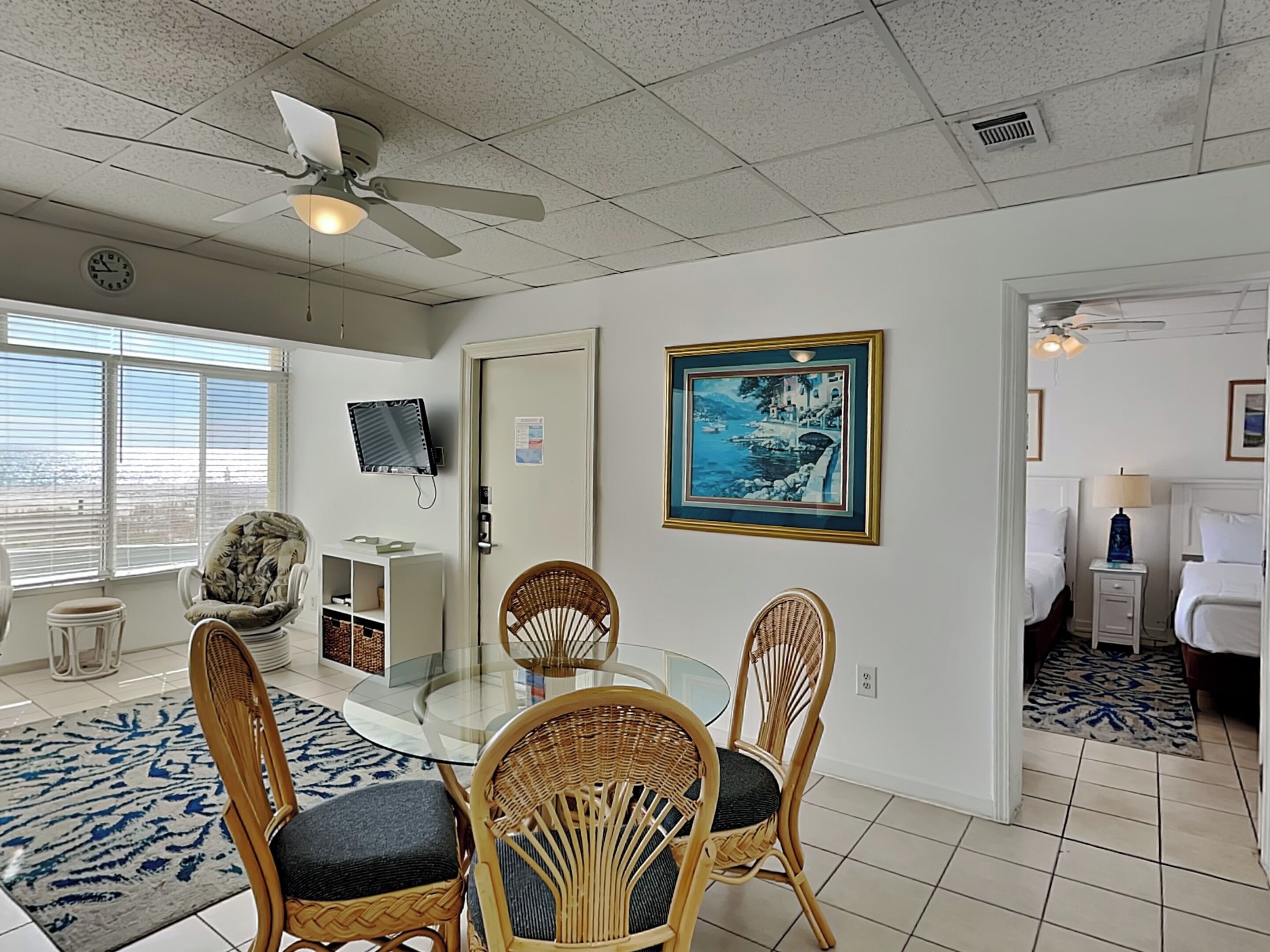A room with a round glass table, two wicker chairs, a ceiling fan, a framed picture, a TV, and a bed with a patterned rug.