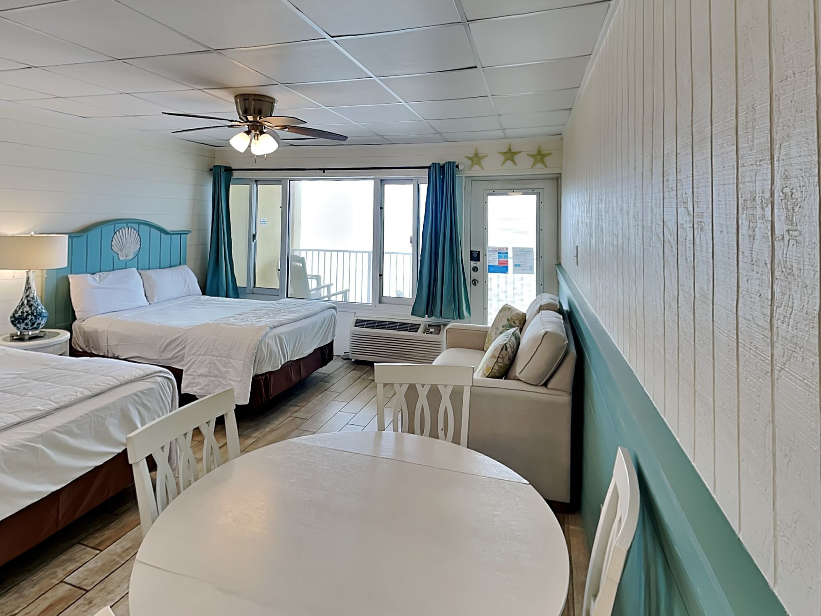 A room with two beds, a ceiling fan, a table, and a view of the balcony.