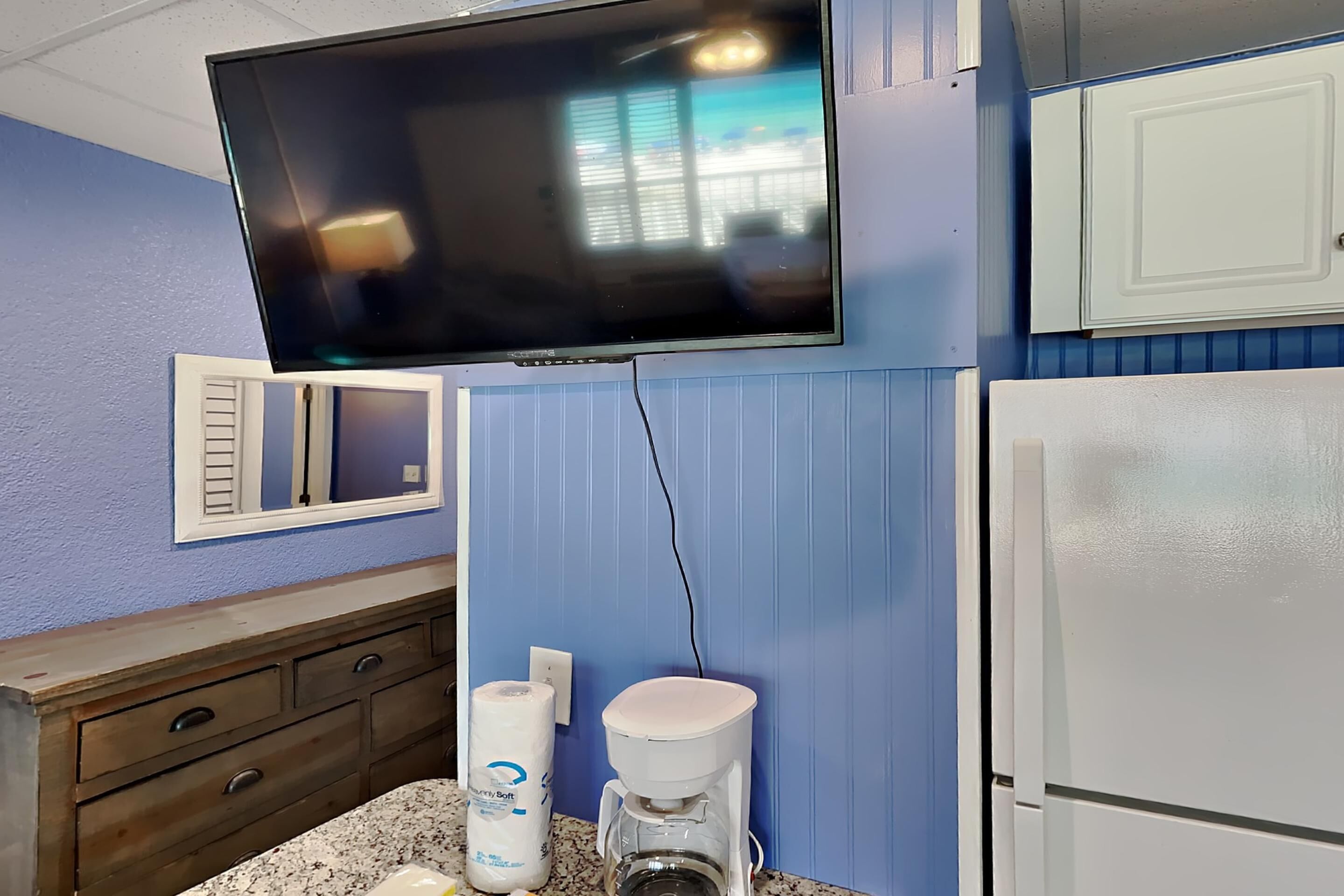 A kitchen with a flat-screen TV mounted above a countertop, a coffee maker, and a refrigerator.