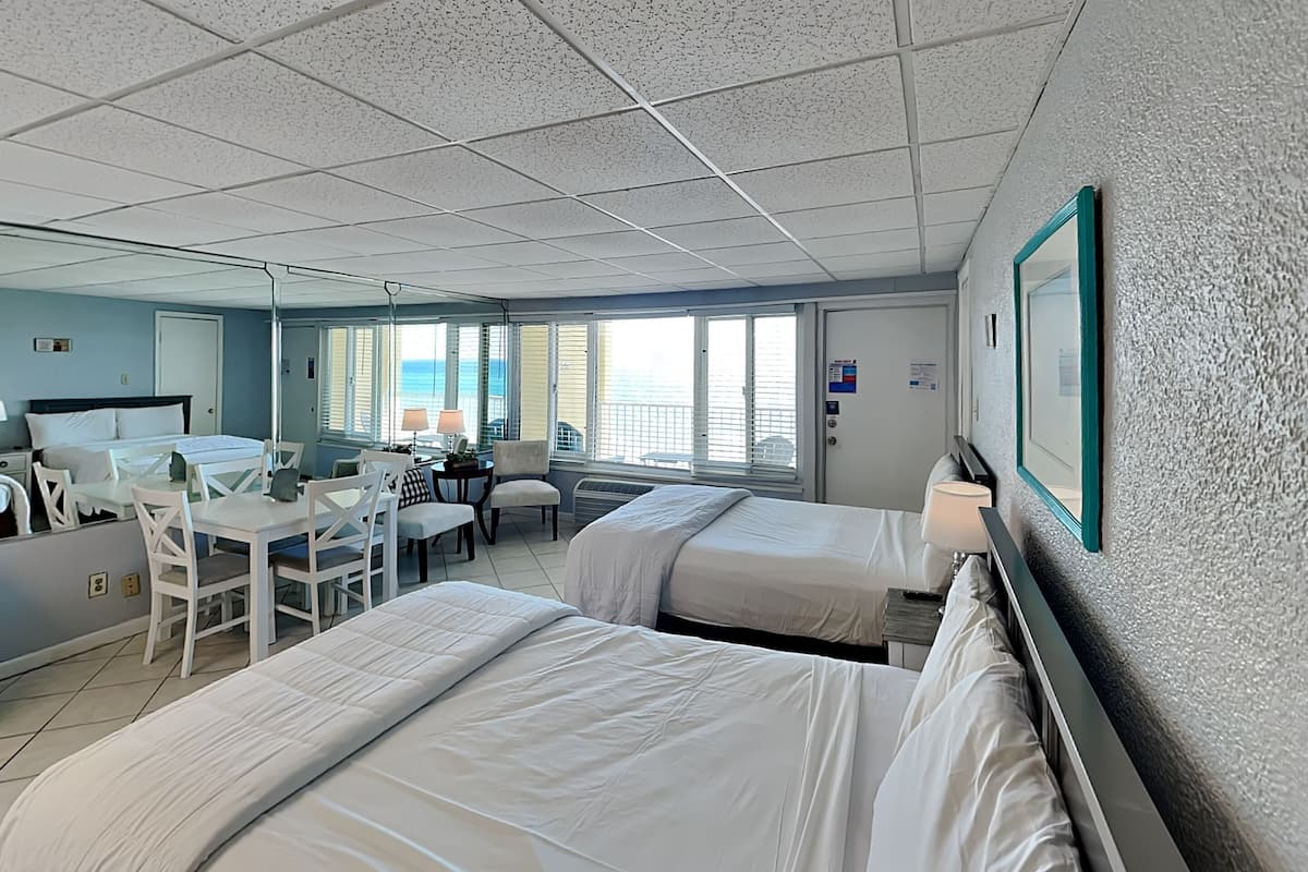 A hotel room with two beds, a desk, chairs, and a view of the ocean.