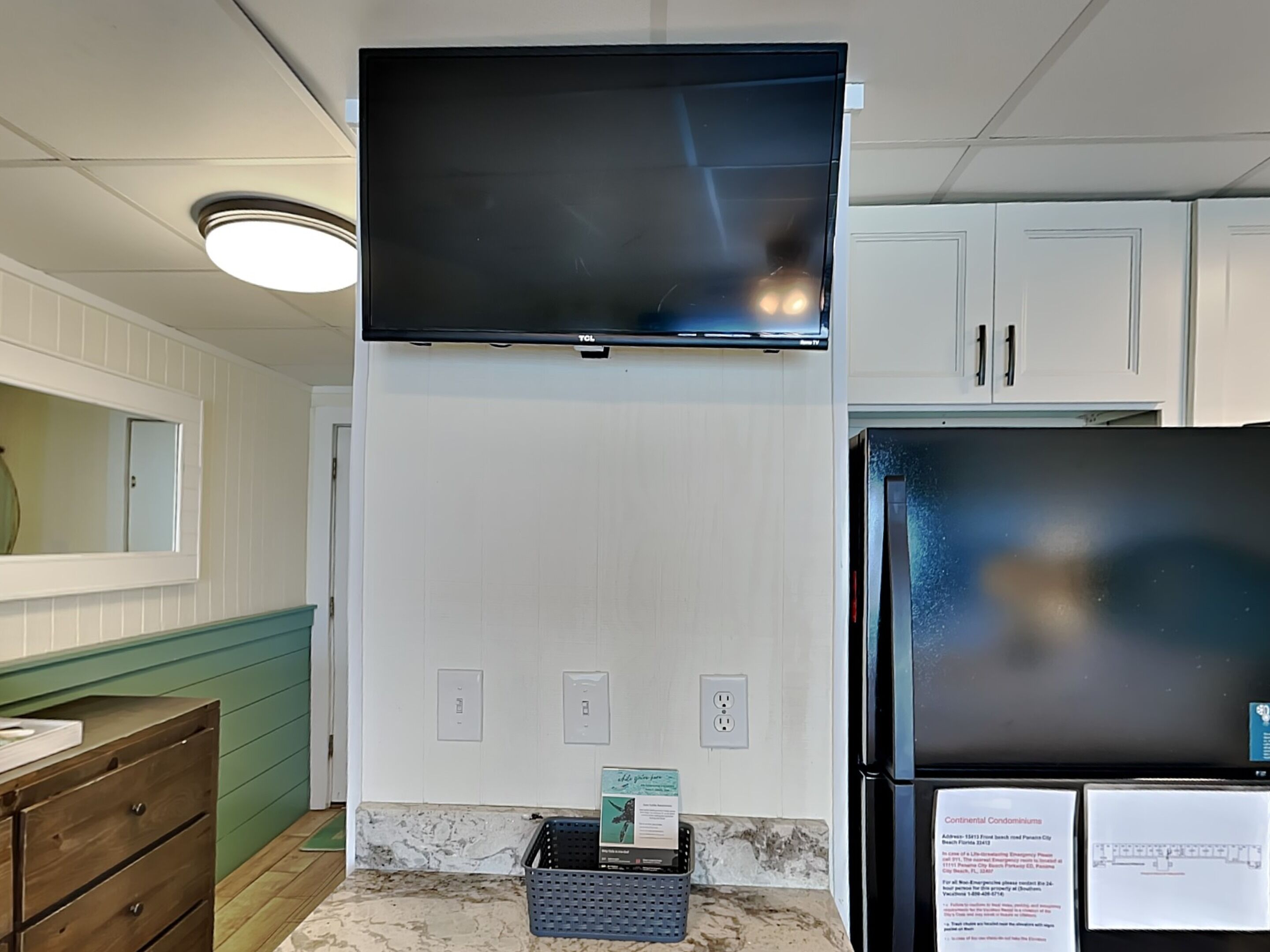 Een keuken met witte kasten, een zwarte koelkast, een wandtelevisie en een houten dressoir.
