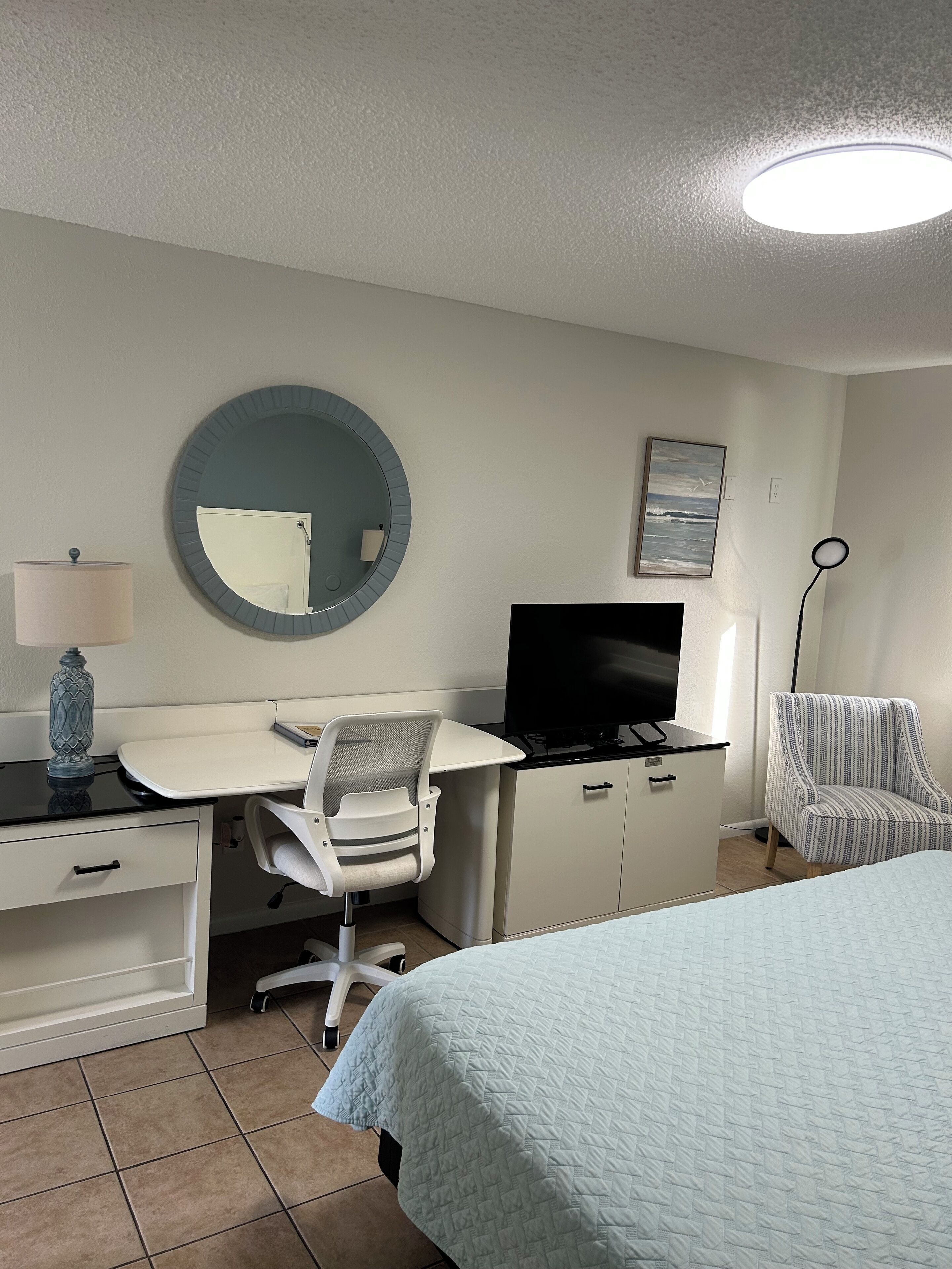 A hotel room with a bed, desk, chair, TV, and a round mirror.