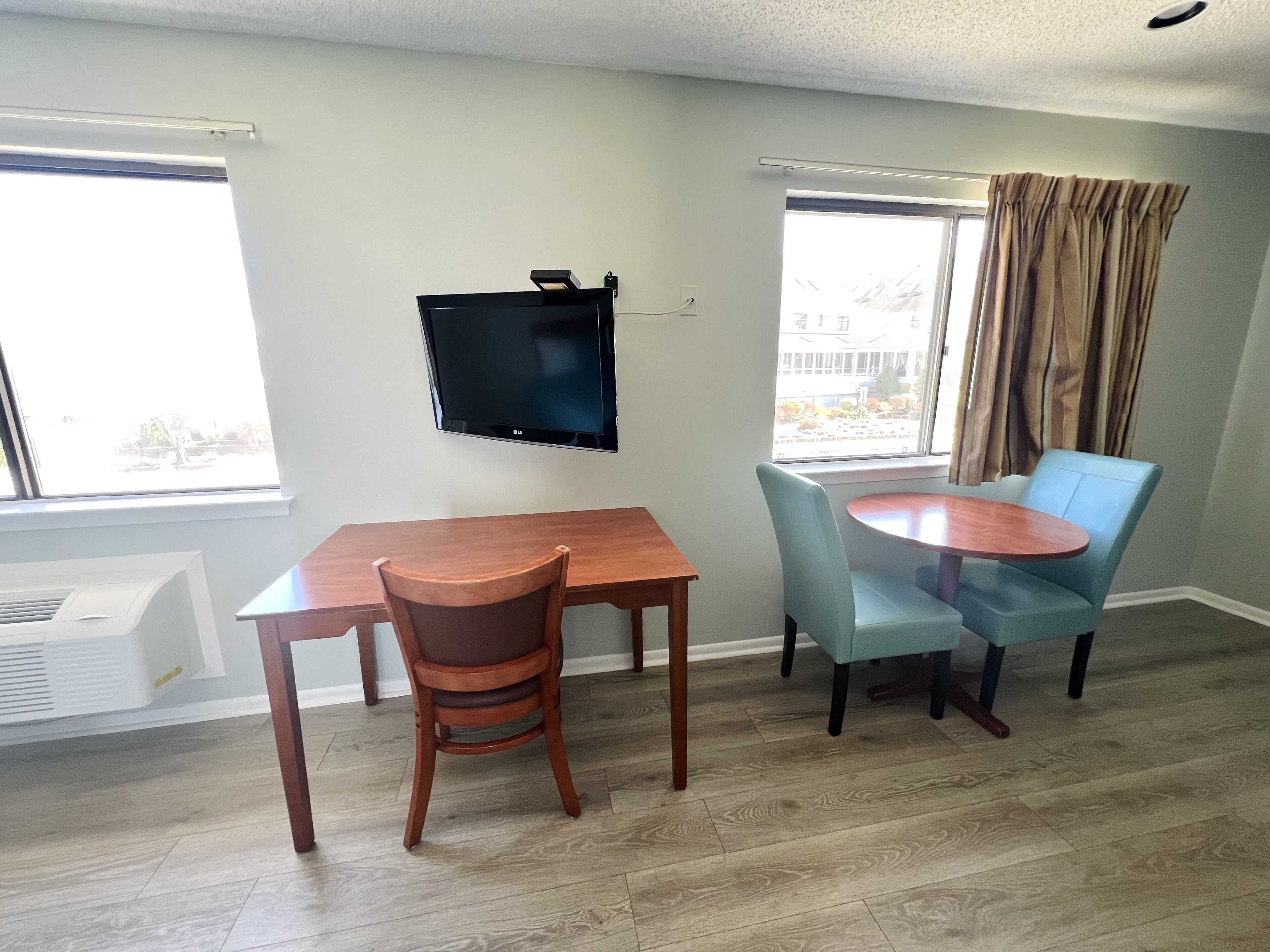 Un comedor con mesa y sillas de madera, un televisor de pantalla plana montado en la pared y ventanas grandes con cortinas.