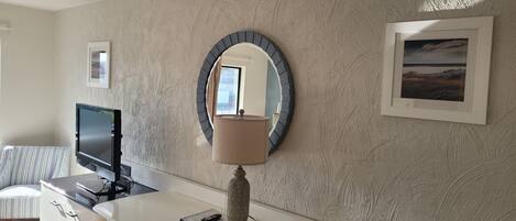 Een hotelkamer met een wit bureau, een televisie, een ronde spiegel, een lamp en een ingelijst schilderij aan de muur.