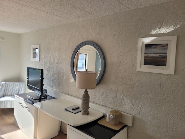 A hotel room with a white desk, a television, a round mirror, a lamp, and a framed picture on the wall.