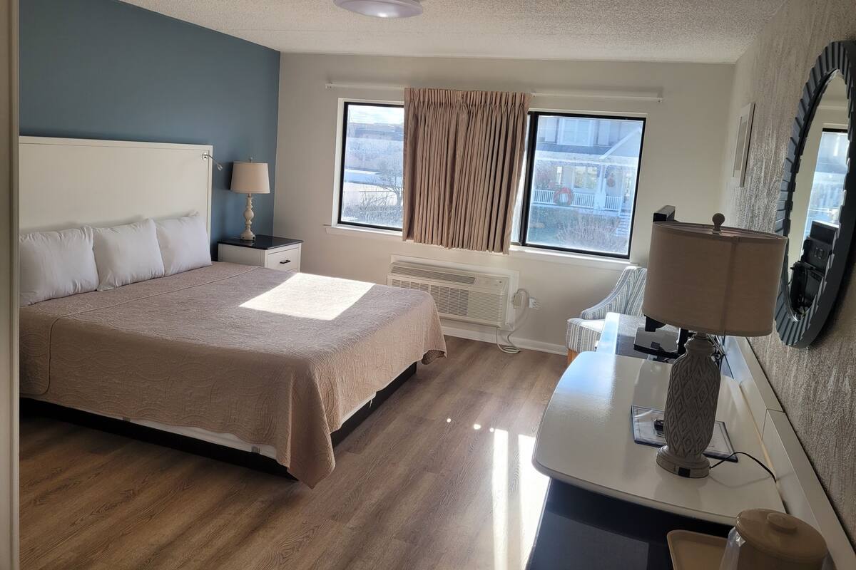 A hotel room with a bed, bedside table, lamp, radiator, and a window with curtains.