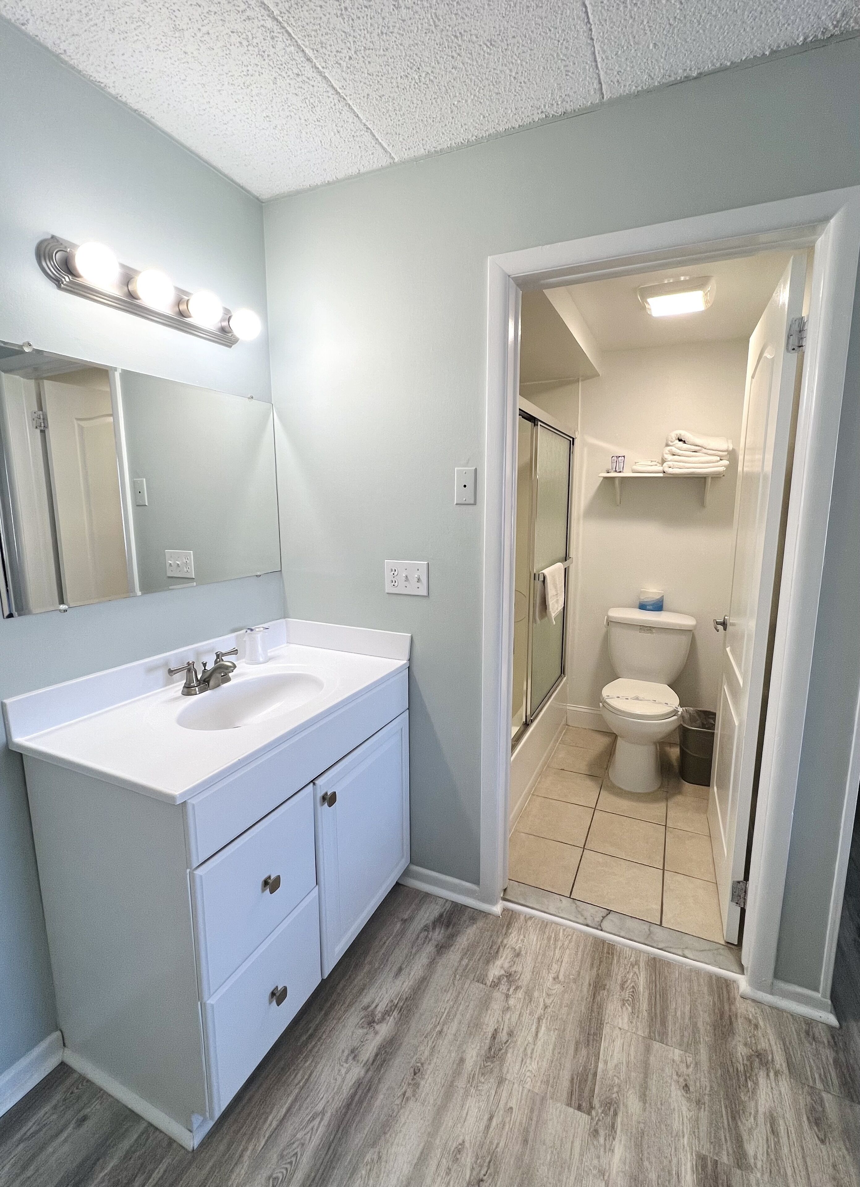 A bathroom with a white vanity, a mirror with lights, a toilet, and a shower area.