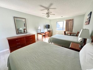 A hotel room with two beds, a desk, a chair, a TV, and a ceiling fan.