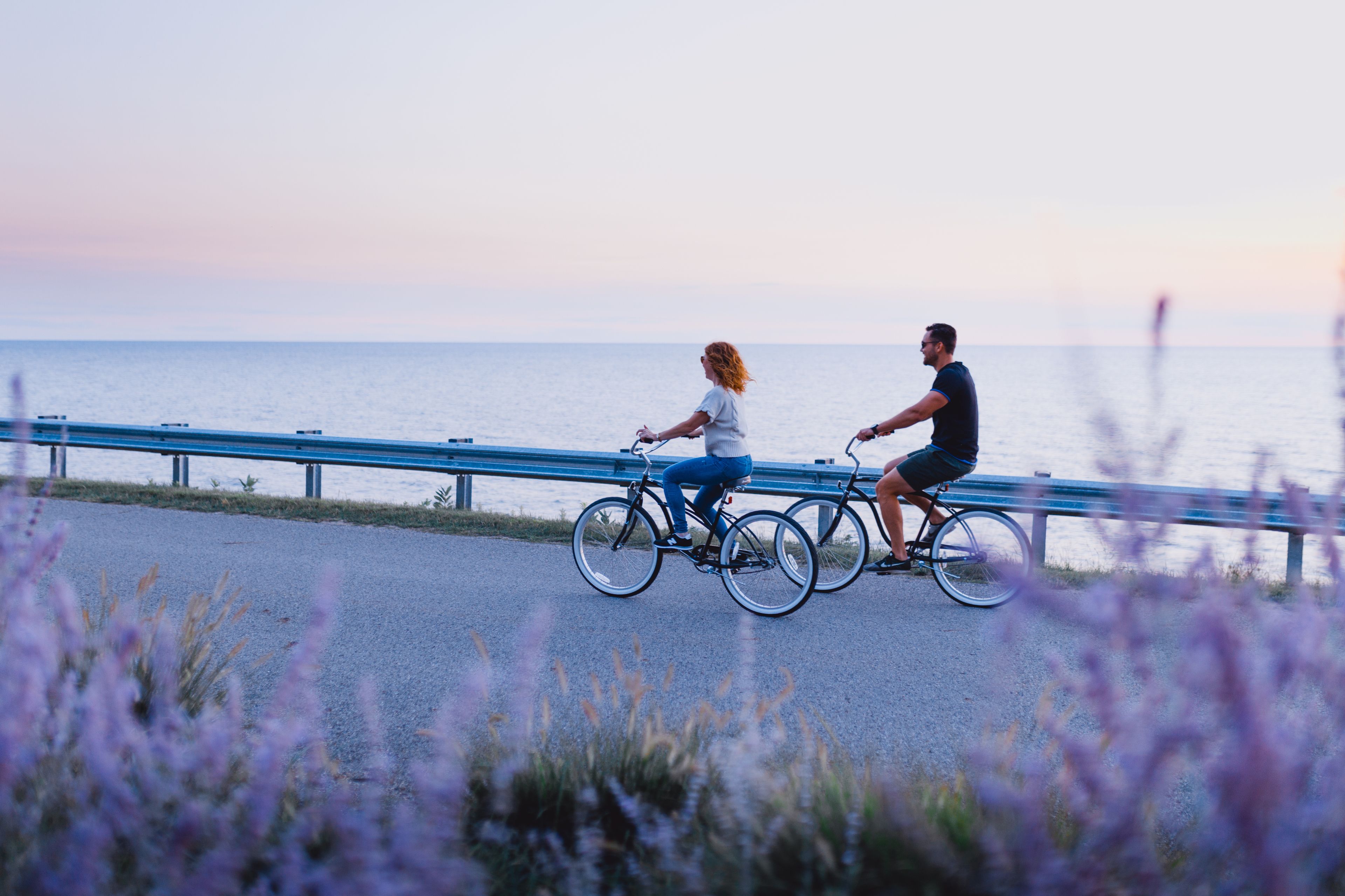 Two people riding bicycles on a paved path beside a body of water.