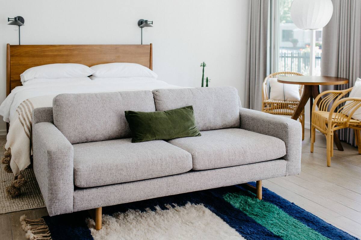 A modern bedroom with a grey sofa, a wooden headboard, a small round table, wicker chairs, and a large window with curtains.