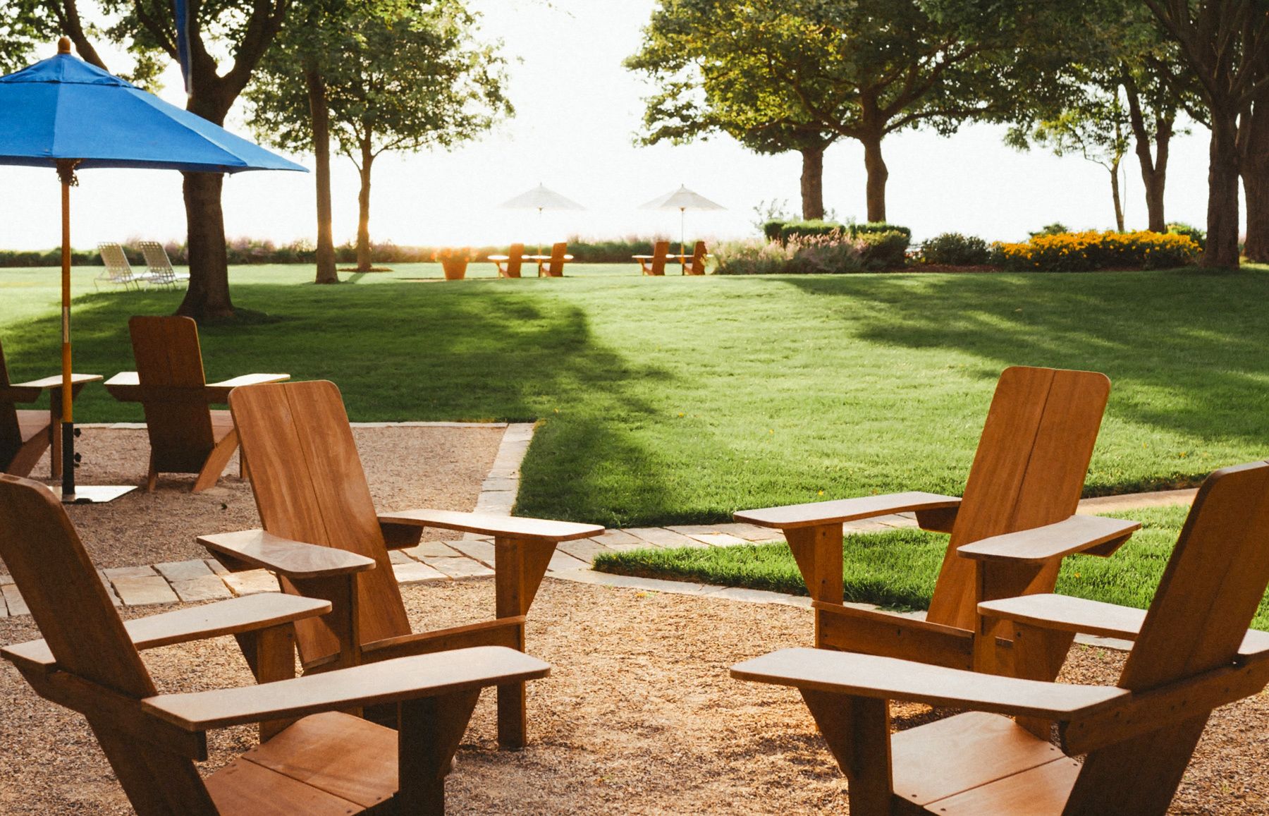 A patio area with wooden chairs and tables, surrounded by a well-maintained lawn and trees.