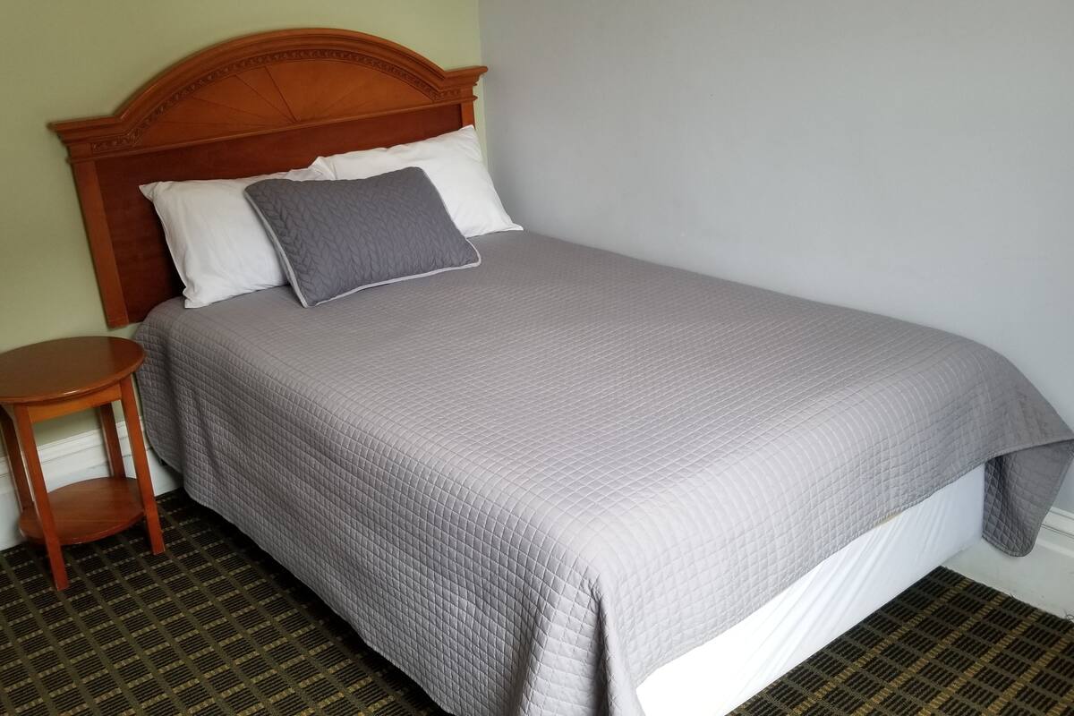 A neatly made bed with a wooden headboard, a small wooden nightstand beside it, and a patterned carpet.