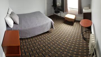 A bedroom with a bed, a wooden nightstand, a small stool, a radiator, and a bathroom with a sink and mirror.