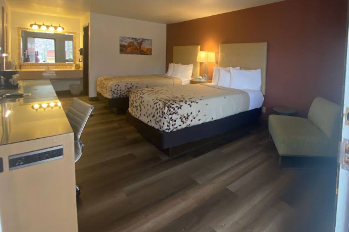 A hotel room with two beds, a desk, a chair, and a small table.