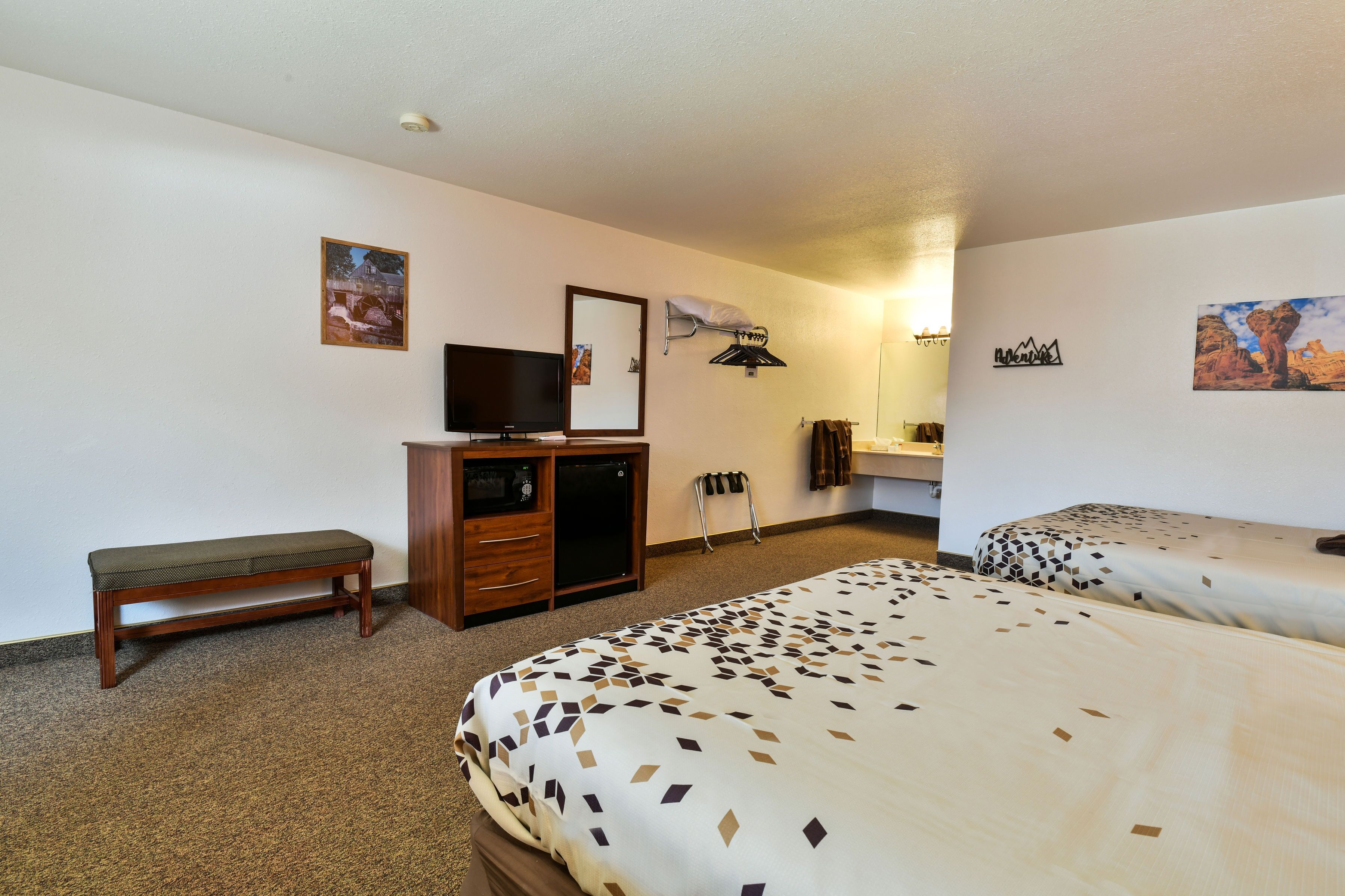 A hotel room with two beds, a bench, a TV, a desk, and a mirror.