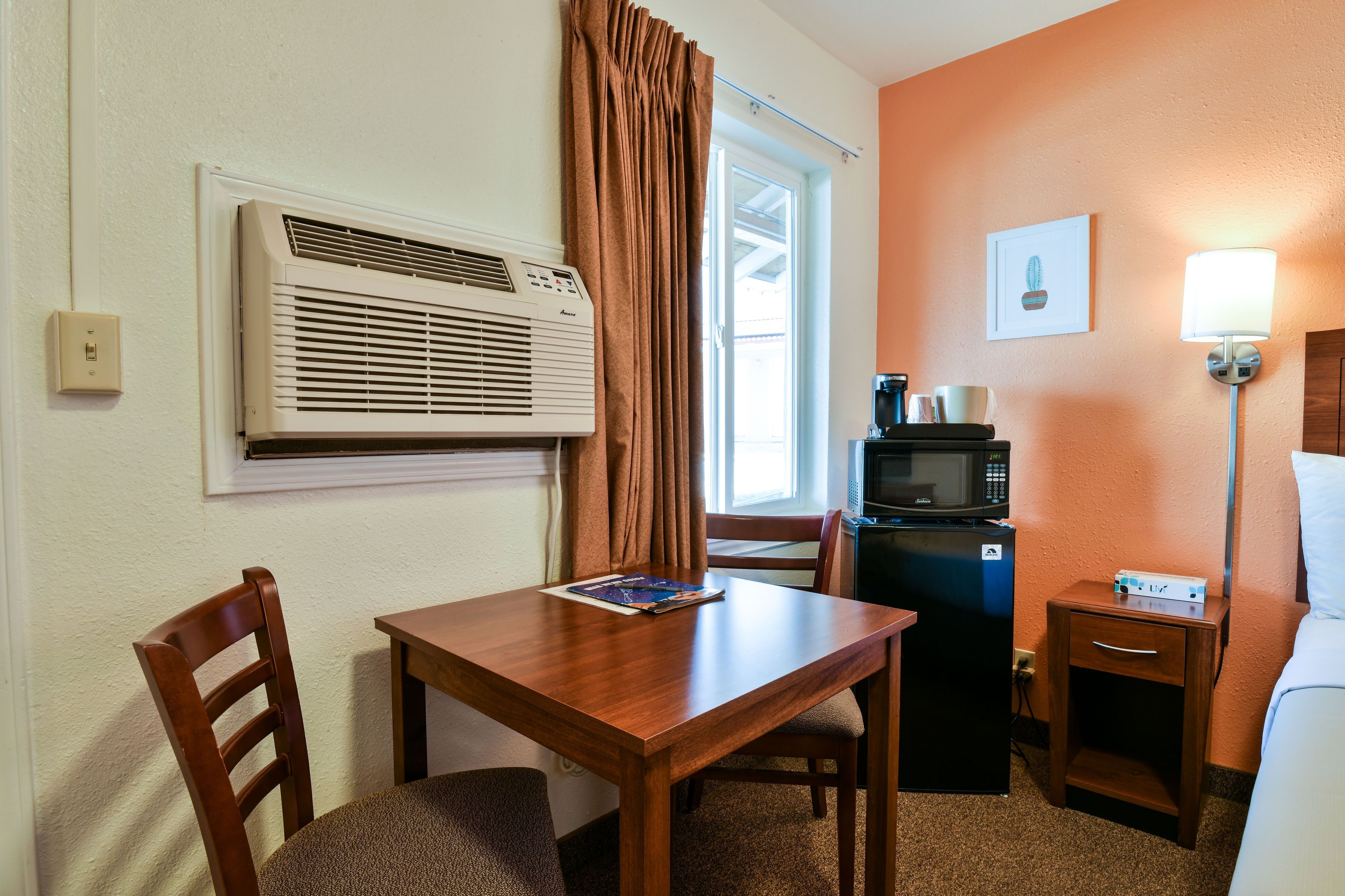 A hotel room with a wooden table, a chair, a microwave, a refrigerator, a lamp, and a window with curtains.