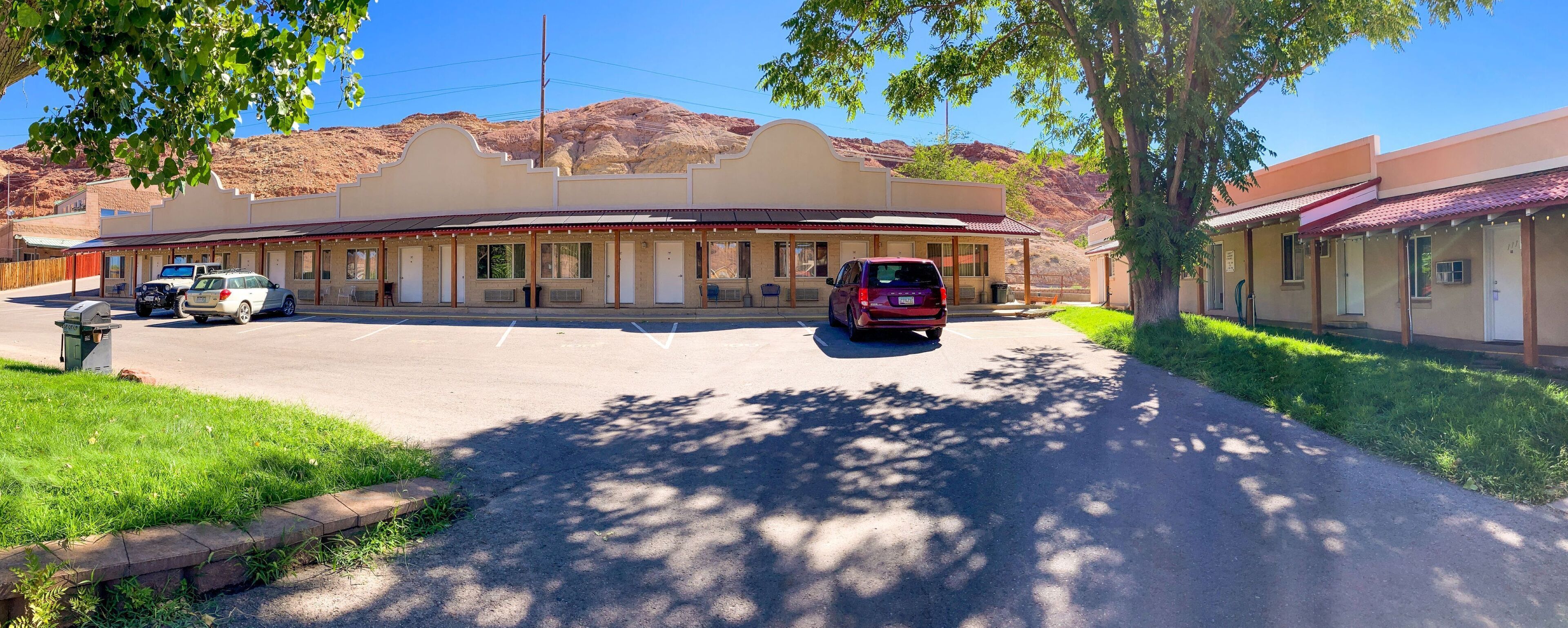 Un motel con un edificio central y dos alas laterales, un estacionamiento con varios coches y montañas al fondo.