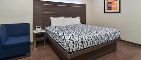 A modern hotel room with a large bed, a blue armchair, a nightstand, and a framed artwork on the wall.