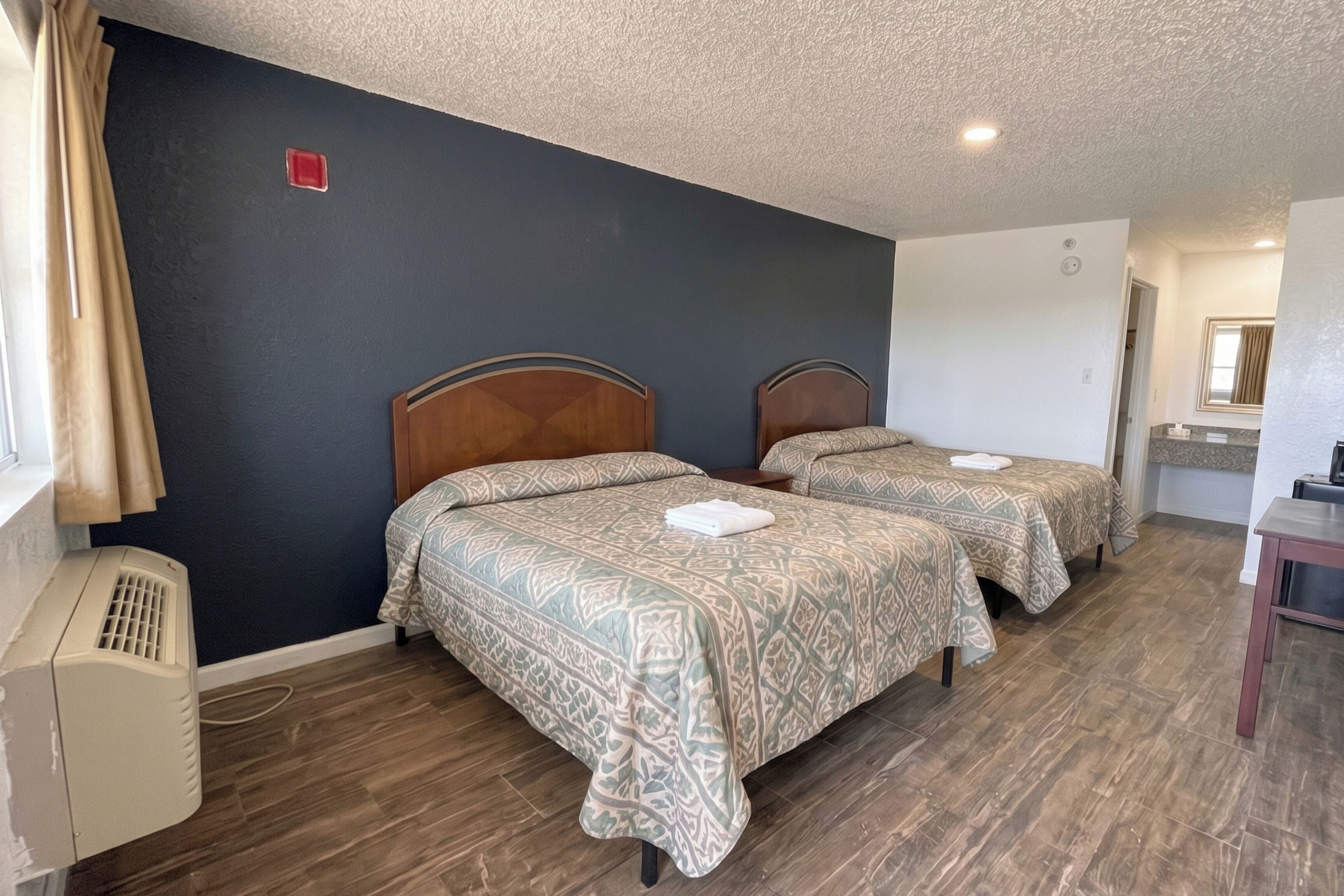 Quarto de hotel com duas camas, um aquecedor, uma janela com cortinas e uma mesa pequena.