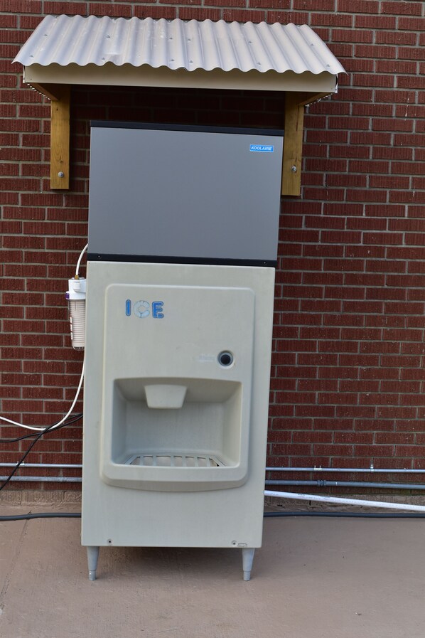 A commercial ice dispenser with a metal roof and a brick wall background.