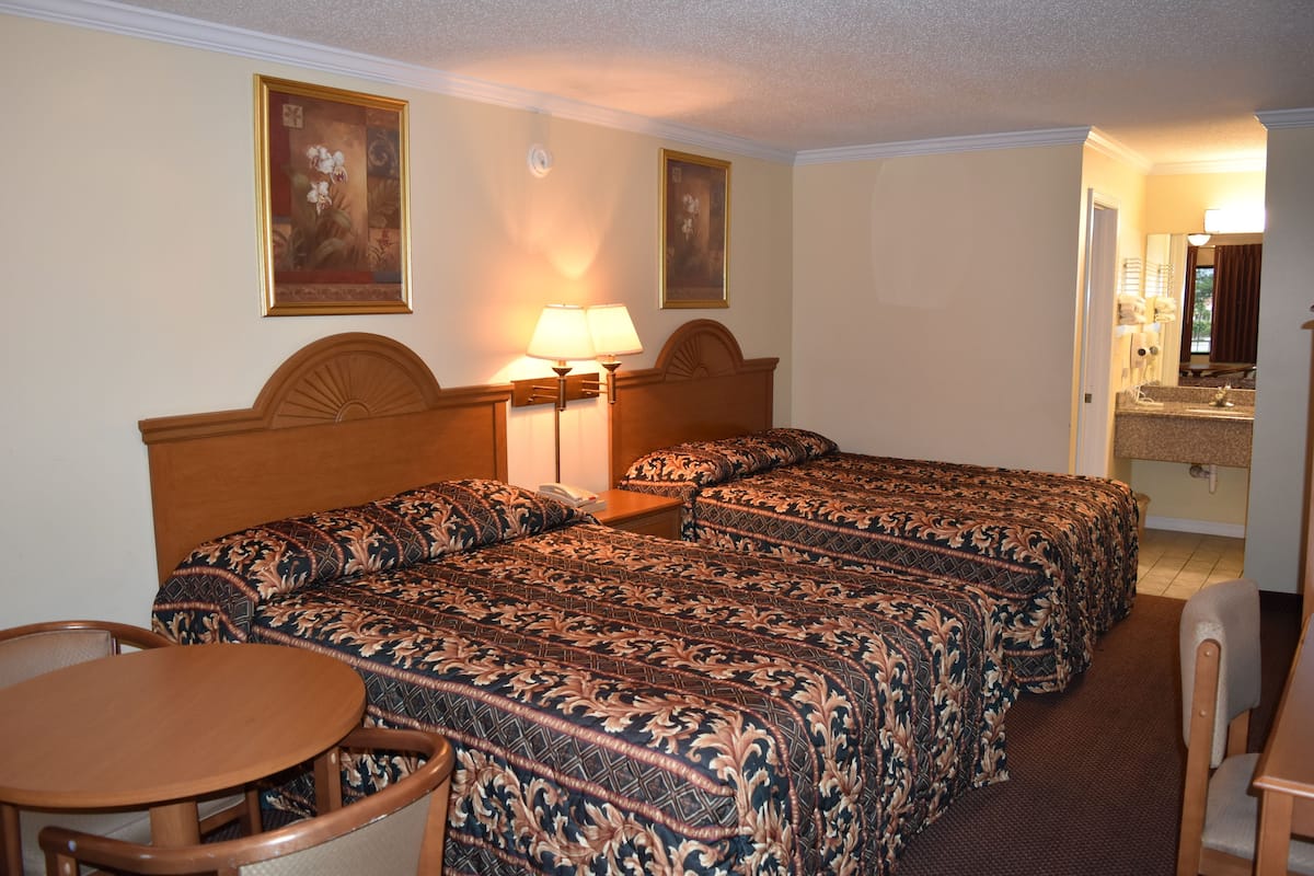 A hotel room with three beds, a desk, and chairs. There are two paintings on the walls.