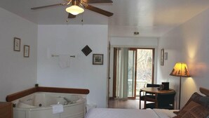 Une chambre avec un grand lit, une baignoire, un ventilateur de plafond, un bureau avec une chaise et une vue à travers une porte vitrée.