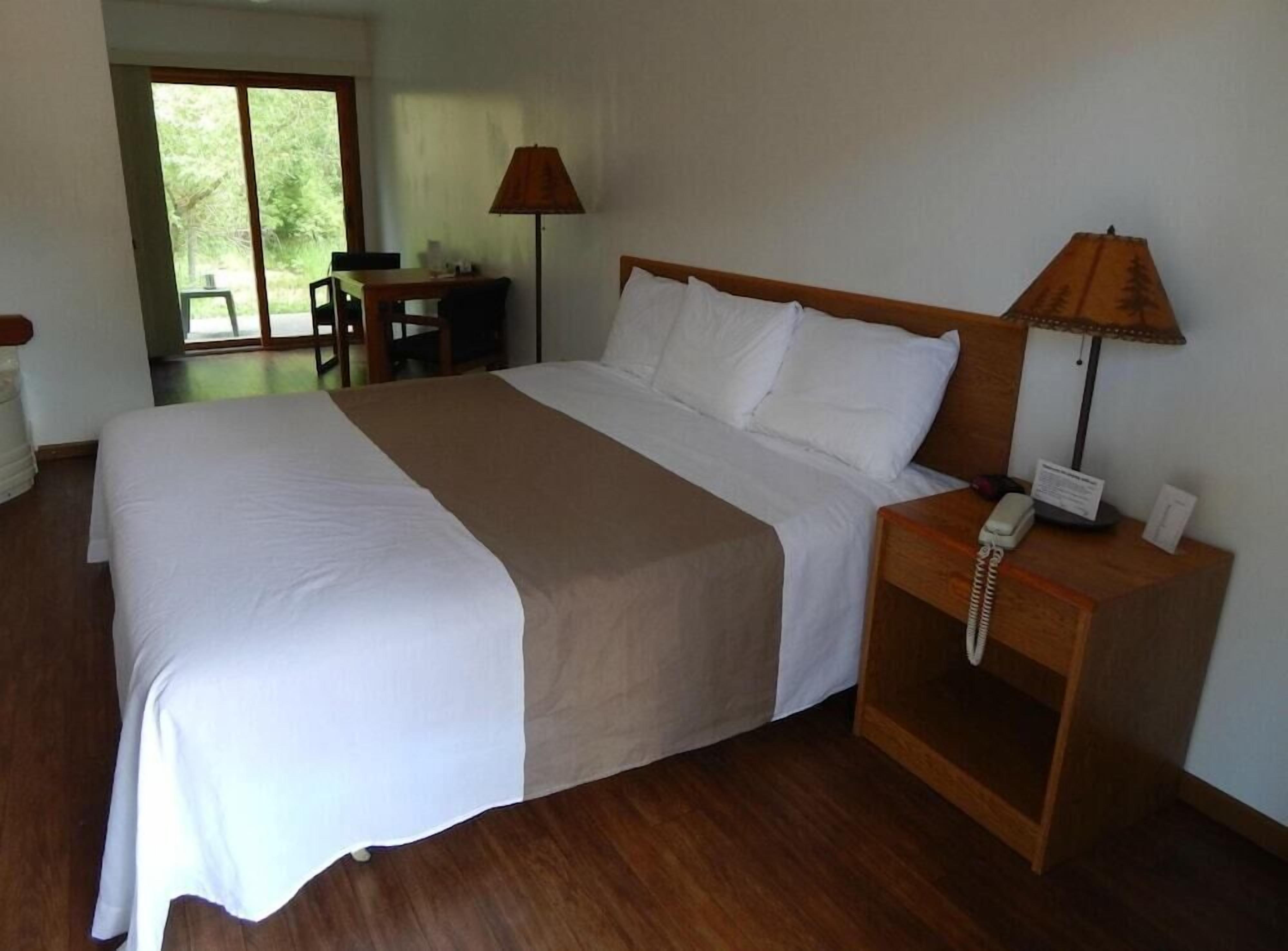 A neatly made bed with white and beige linens, a wooden nightstand with a telephone, and a desk with a lamp.