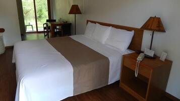 A neatly made bed with white and beige linens, a wooden nightstand with a telephone, and a desk with a lamp.