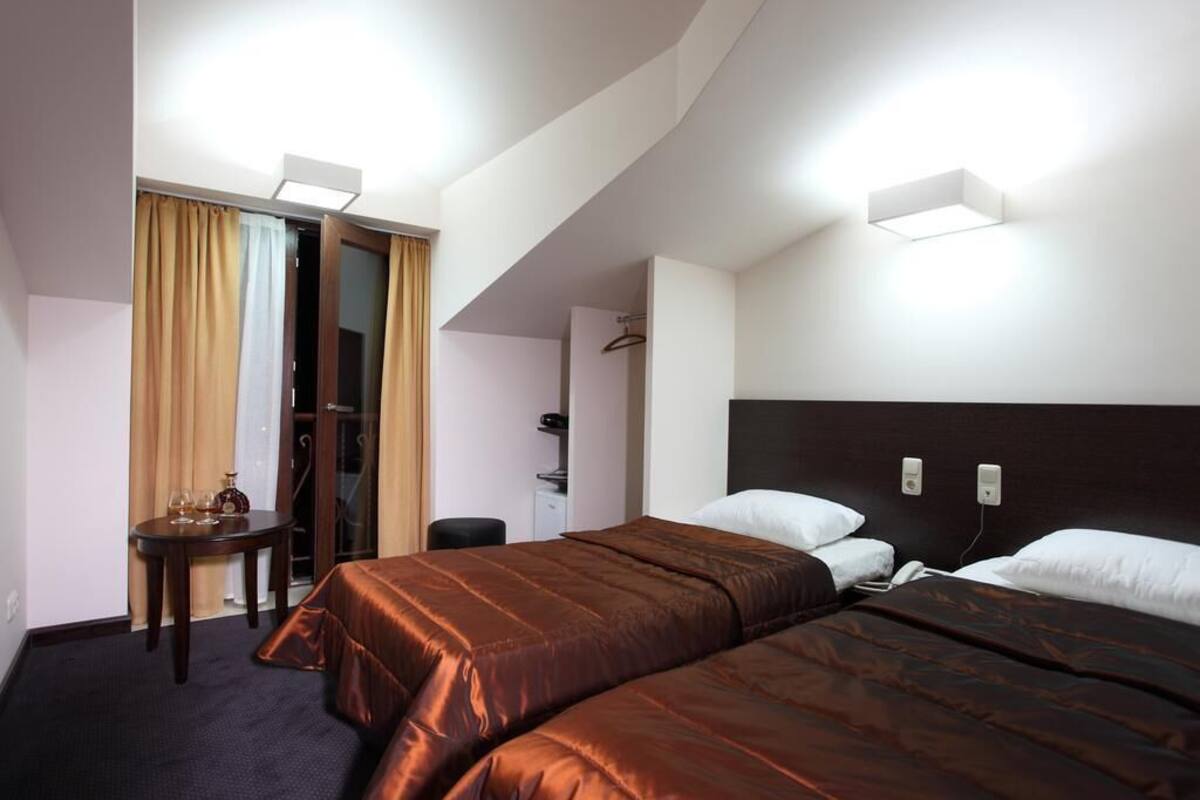 Habitación de hotel con dos camas, un escritorio con silla, una mesita con una botella y vasos, y un armario.
