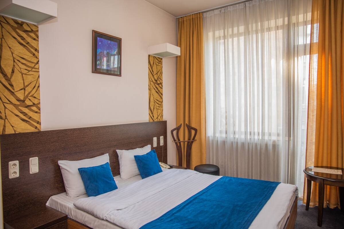 Habitación de hotel con una cama grande, cabecera de madera, mesita de noche, una silla y una ventana con cortinas.
