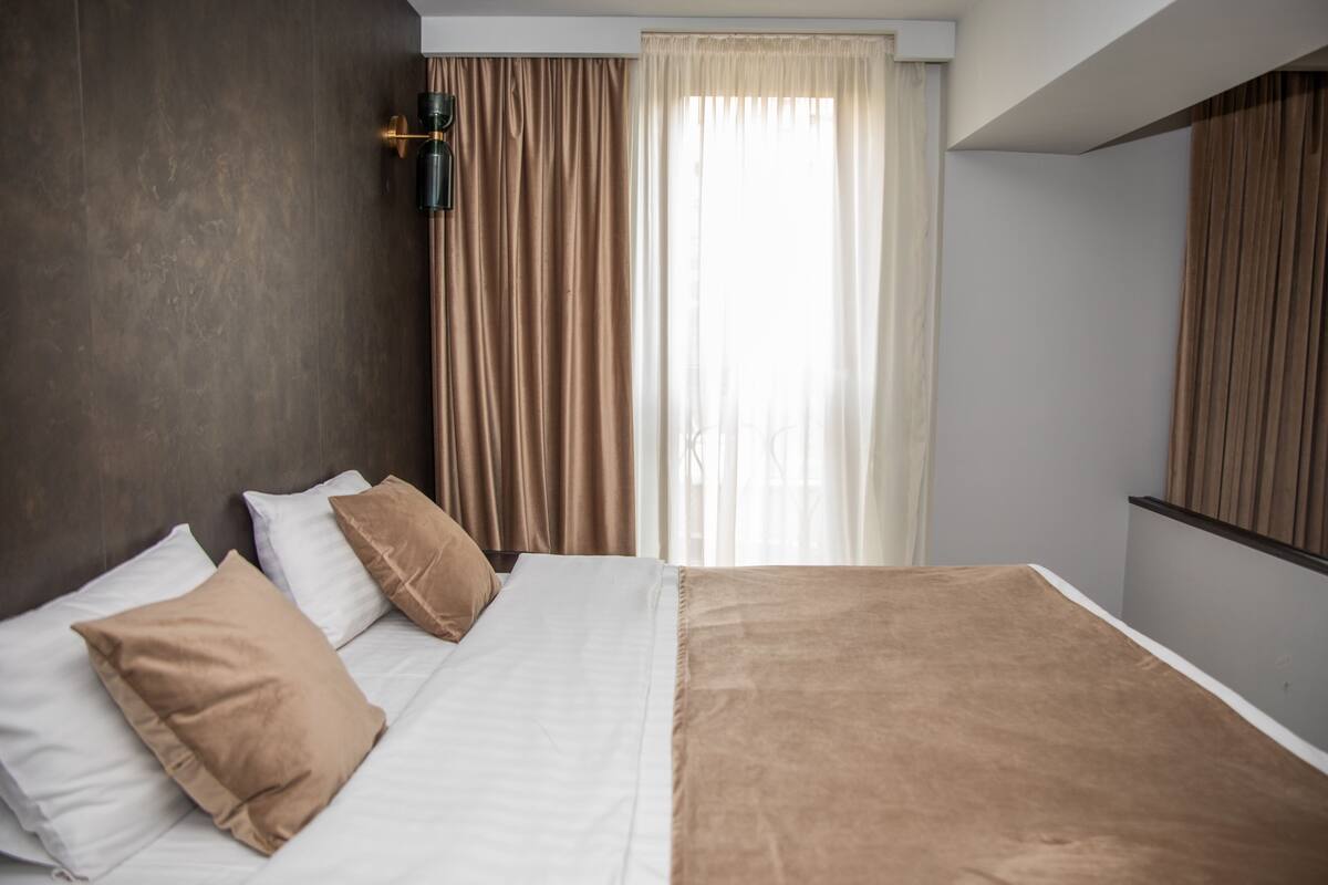 Una habitación de hotel con una cama grande, ropa de cama marrón y blanca, una pared oscura, una ventana con cortinas y una lámpara fijada a la pared.