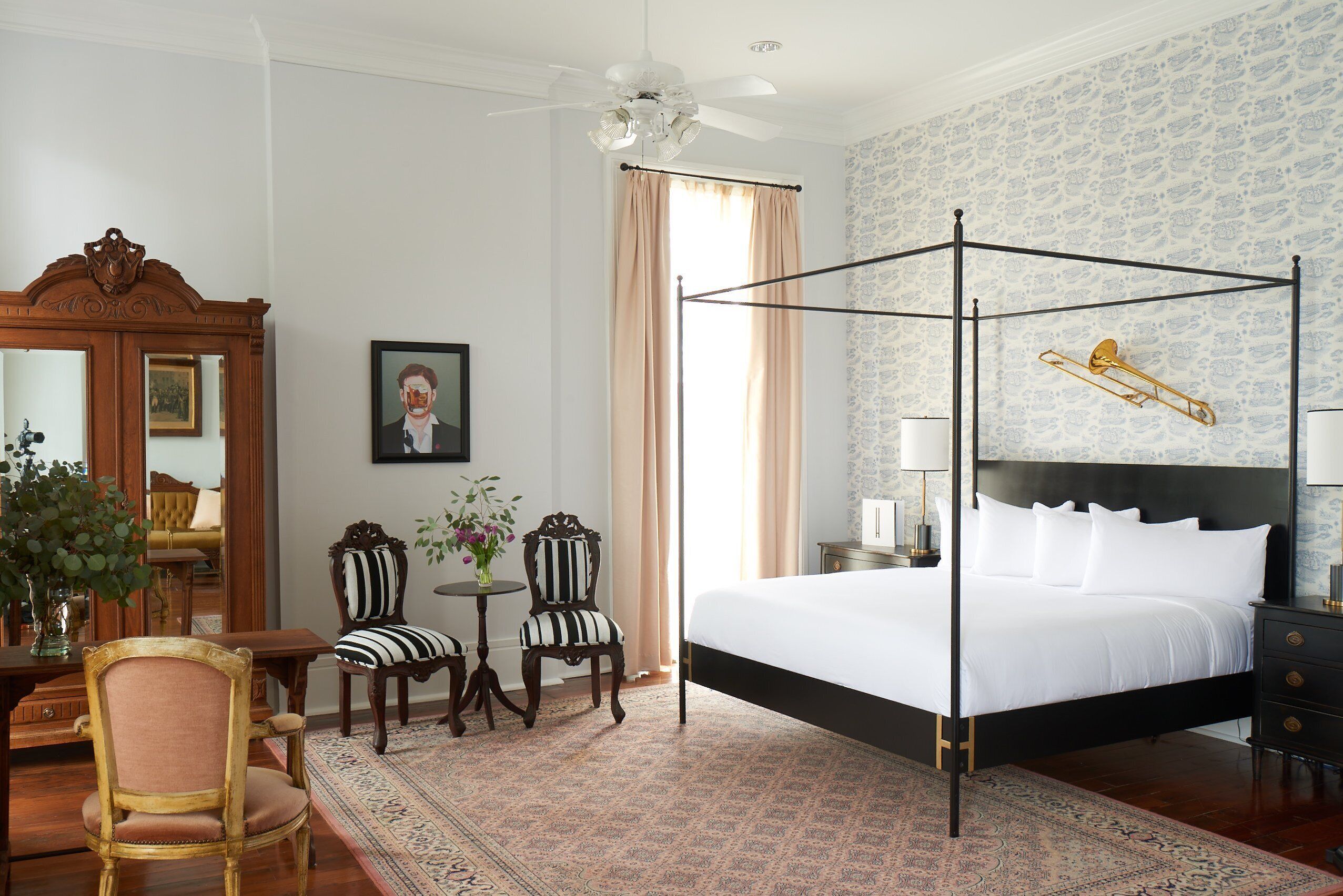 A four-poster bed with a white bedspread, a round table with chairs, a mirror, and a patterned rug.