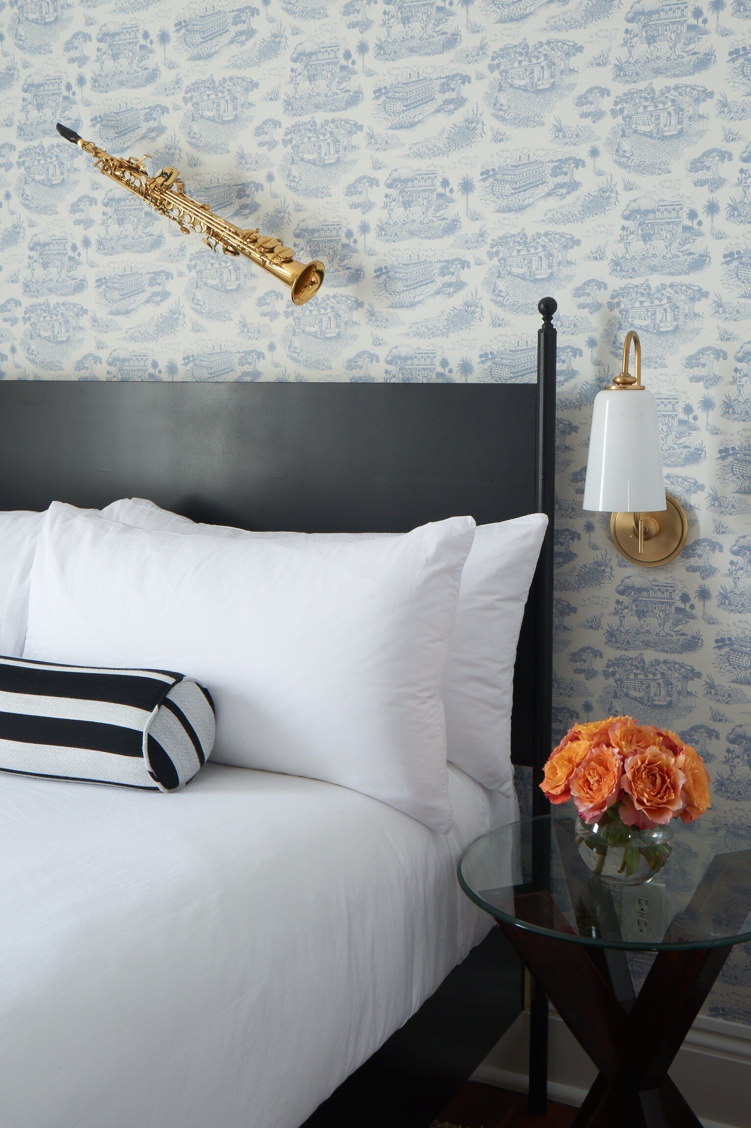Una cama con ropa blanca, un cabecero negro y una almohada a rayas. Hay una mesita de cristal con un jarrón de flores.