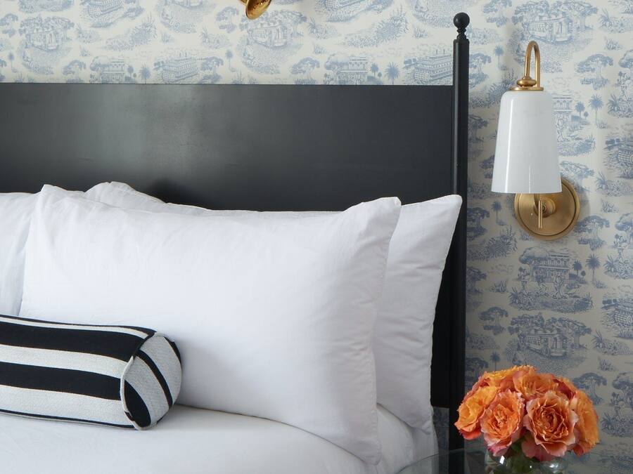 Ein Bett mit weißer Bettwäsche, einem schwarzen Kopfteil und einem gestreiften Kissen. Ein Glastisch mit einer Blumenvase steht daneben.