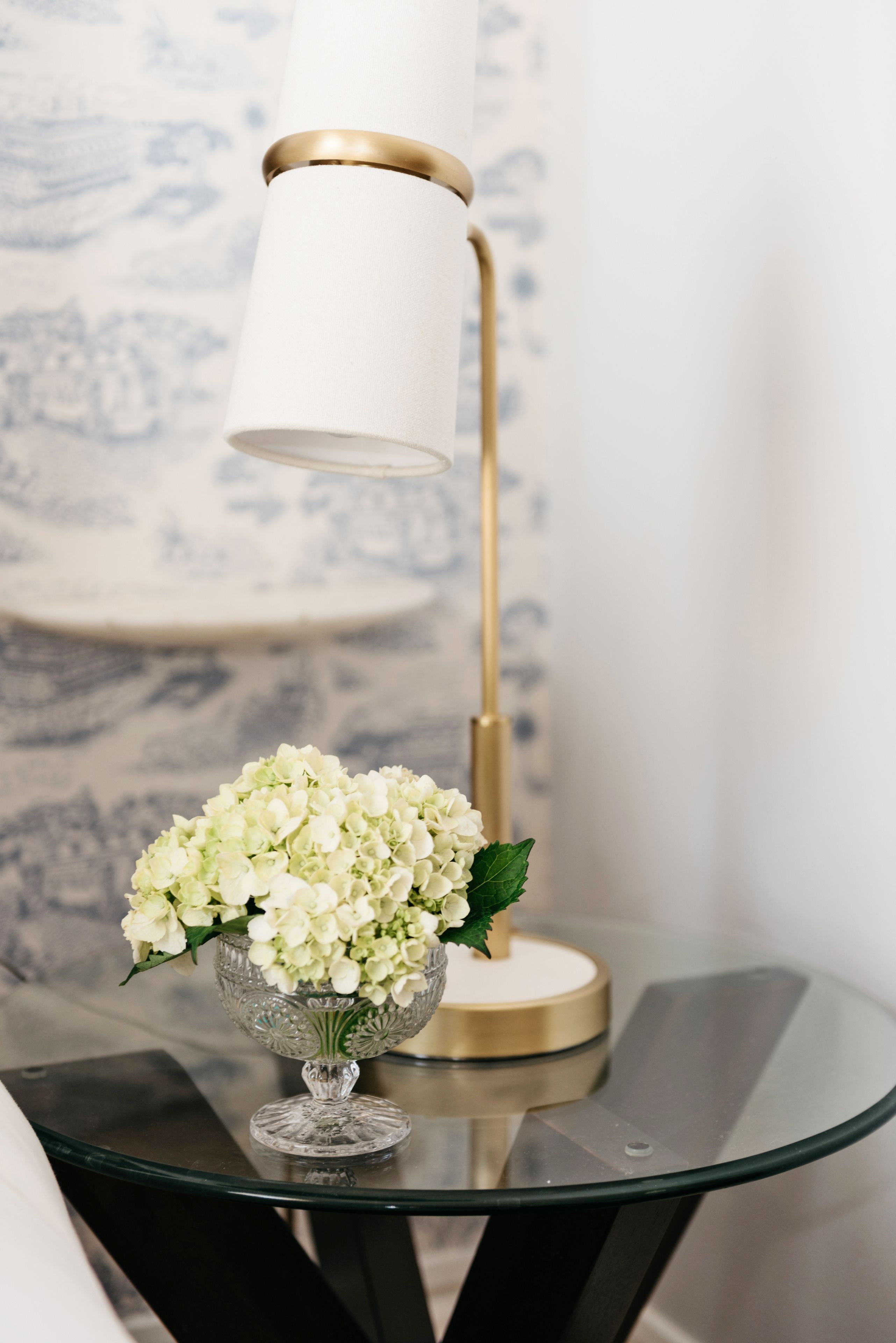 Ett bord med en glasskiva, en guldfärgad lampa och en glasskål fylld med vita blommor.