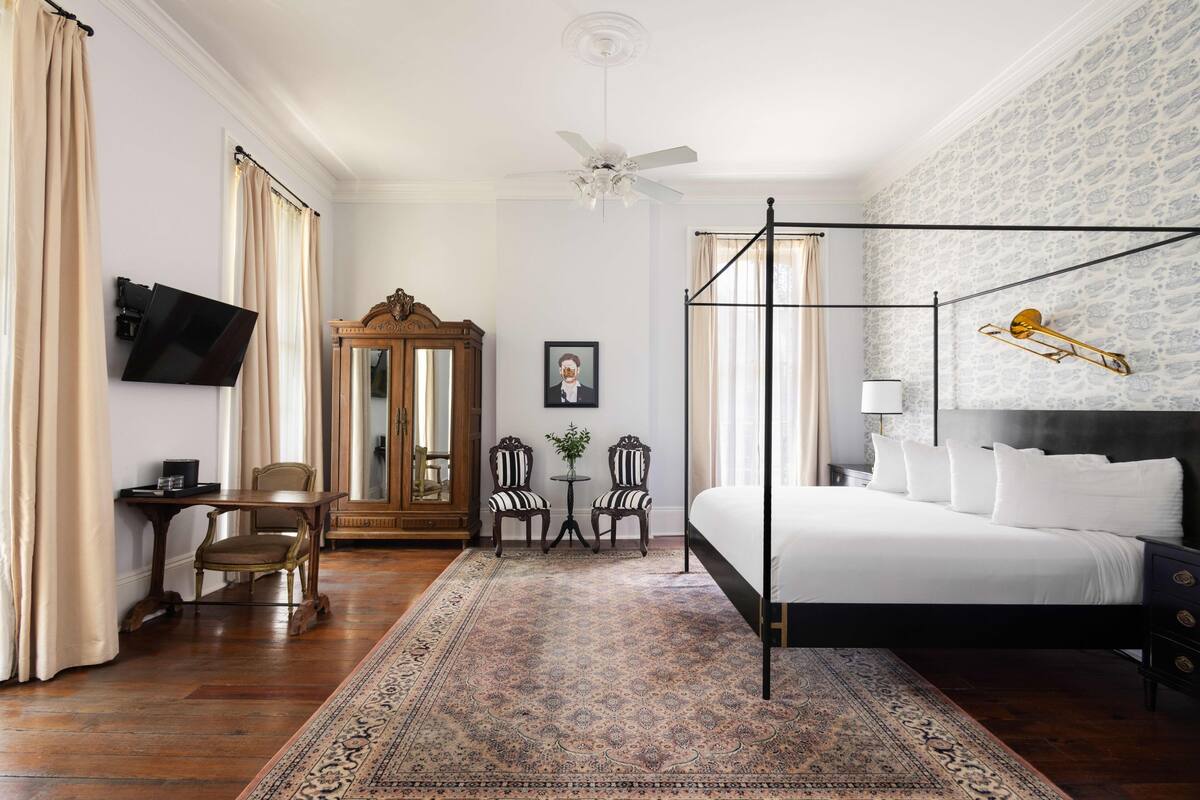 A hotel room with a four-poster bed, a large mirror, a television, a small table with chairs, and a patterned rug.