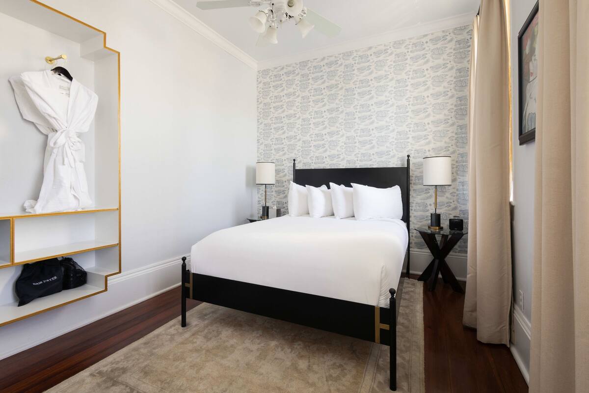 A neatly arranged hotel room with a bed, bedside lamps, a robe, and a shelf with a bag.