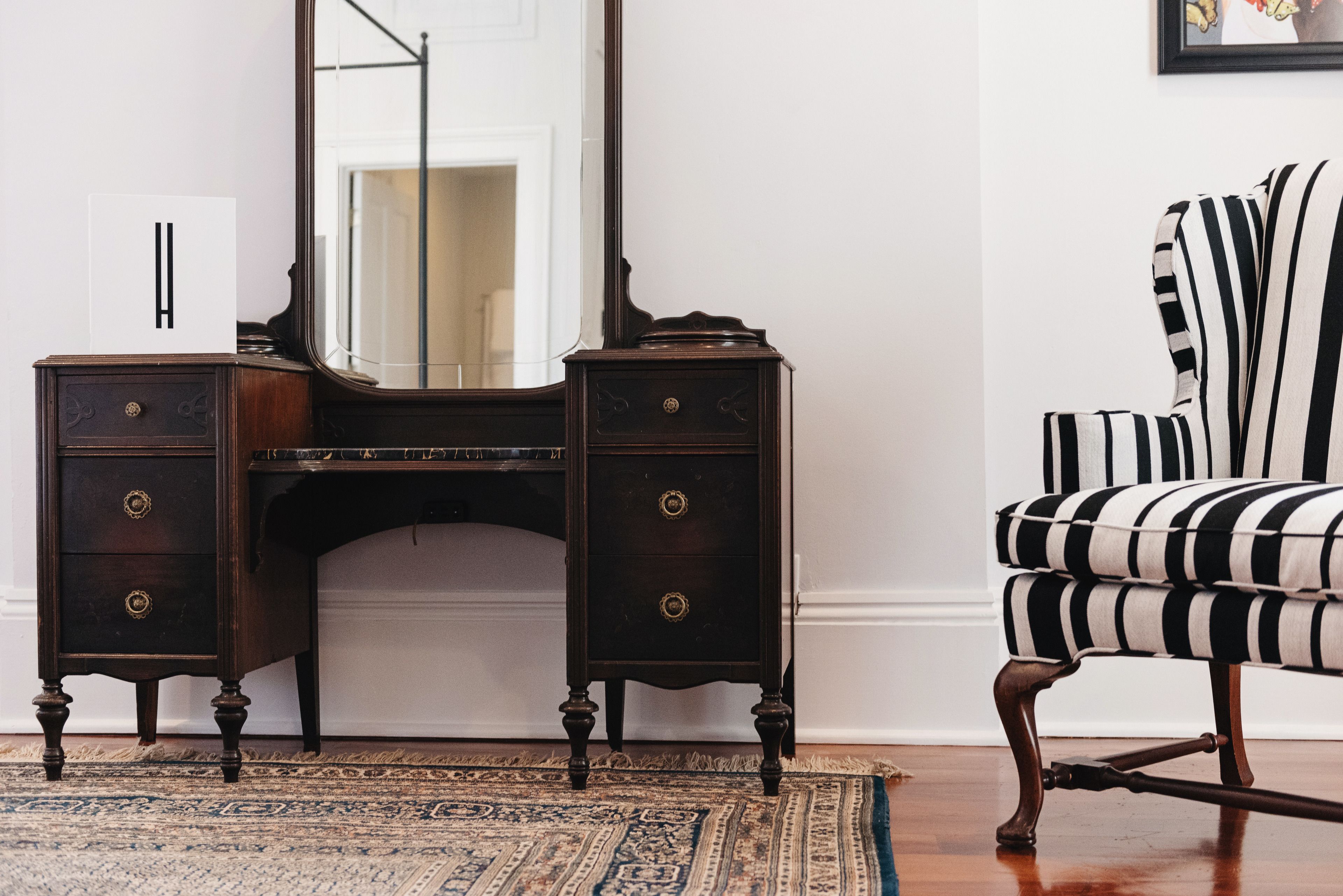 A dark wooden desk with brass handles, a large mirror, and a striped chair.