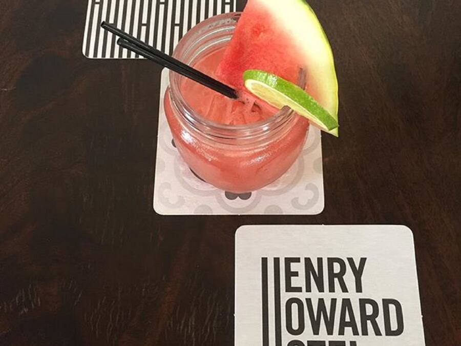 A glass of pink drink with a slice of watermelon and a lime wedge, placed on a coaster with a card reading "ENRY HOWARD HOTEL" underneath.