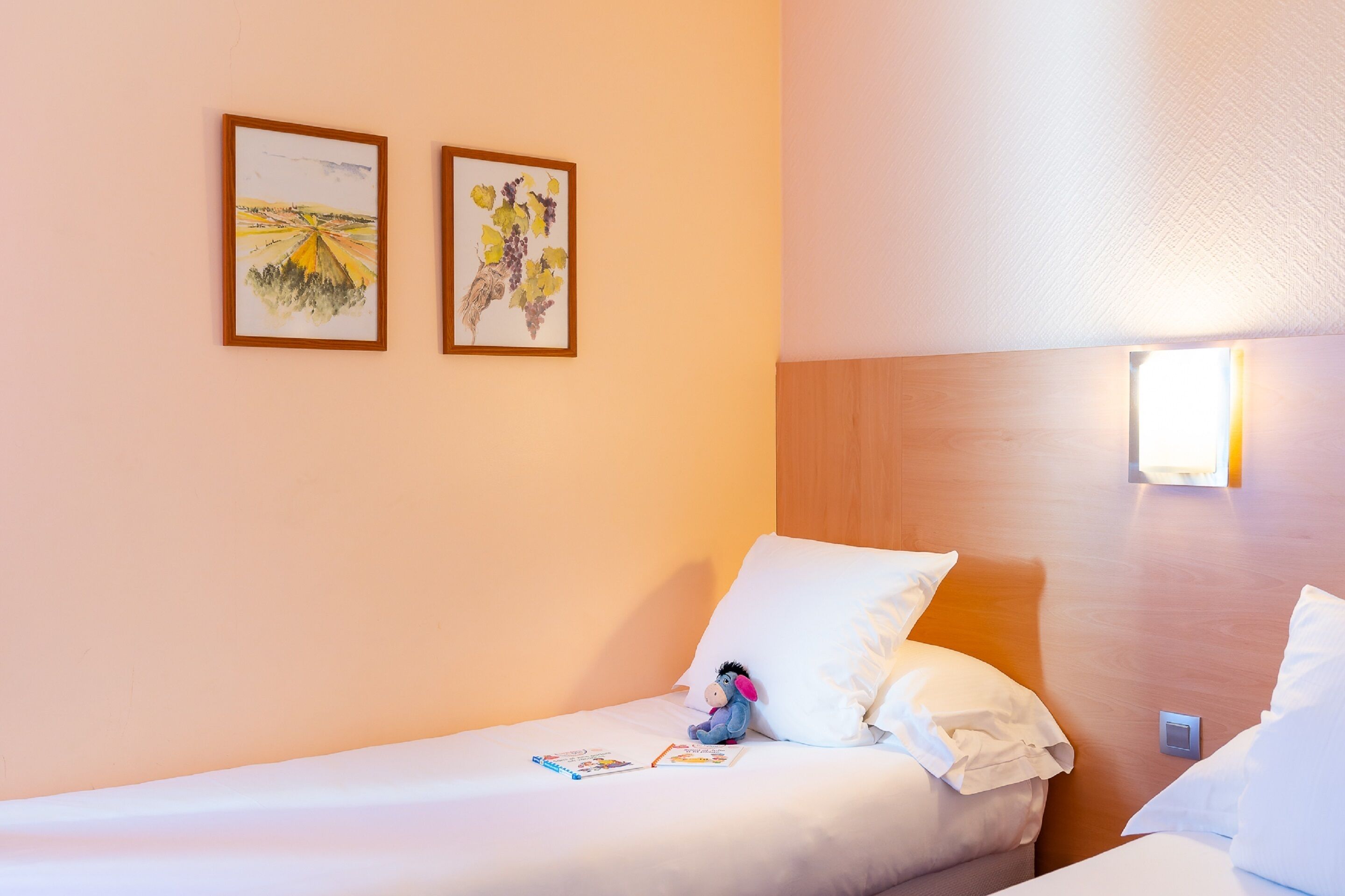 Une chambre d’hôtel avec un lit simple, une table de chevet, deux œuvres d’art encadrées accrochées au mur et un luminaire fixé au mur.
