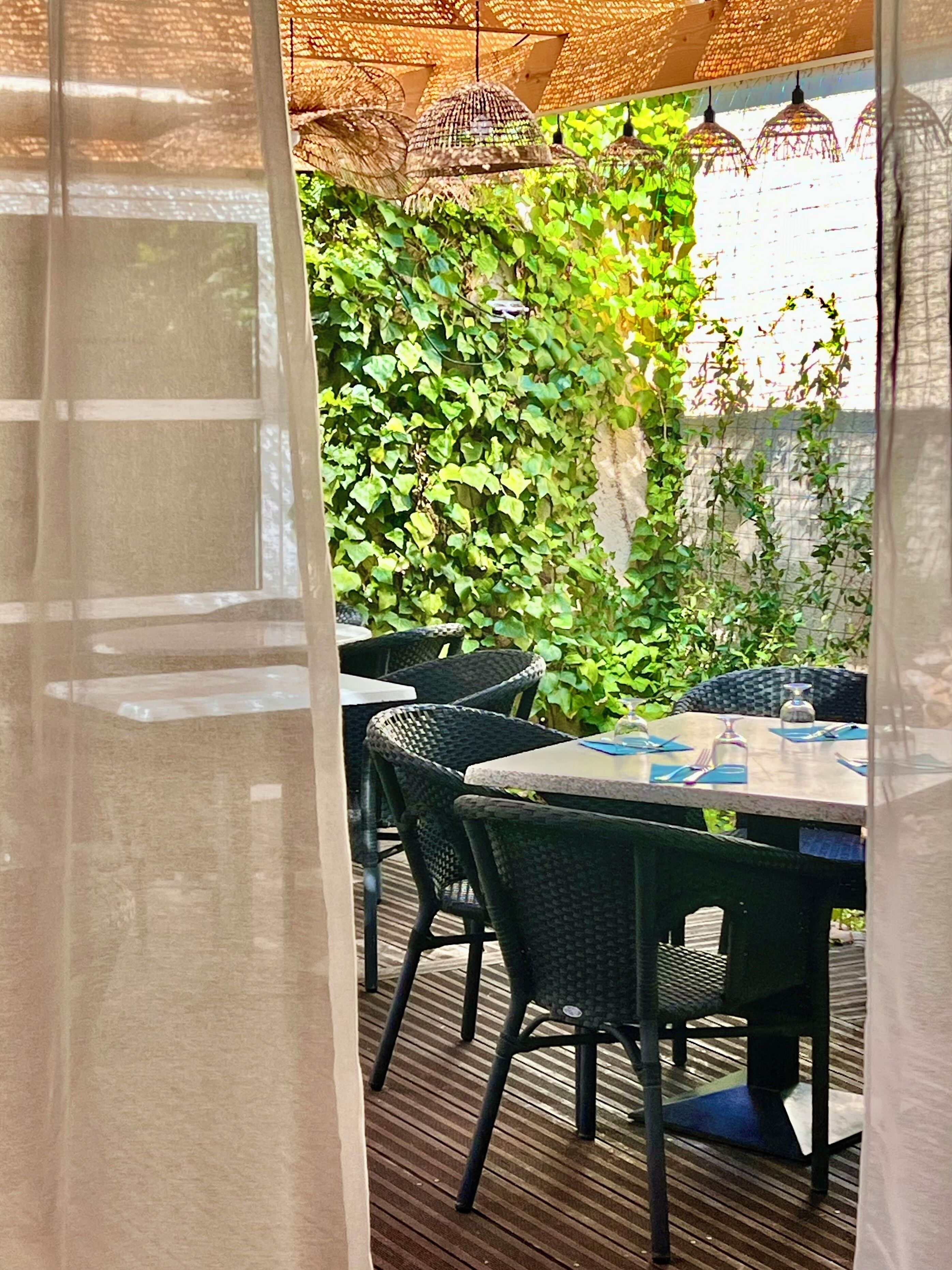 An outdoor dining area with wicker chairs and a table set for two, surrounded by lush green foliage.