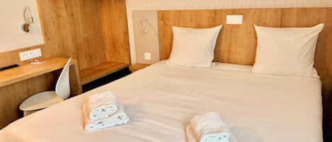 Una habitación de hotel con cabecera de madera, dos almohadas, un escritorio con silla y un perchero para toallas.