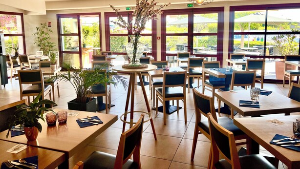 Een restaurant met meerdere tafels en stoelen, grote ramen en hangende lampen.