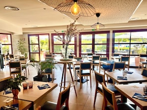 Un restaurante con varias mesas y sillas, amplias ventanas y lámparas colgantes.