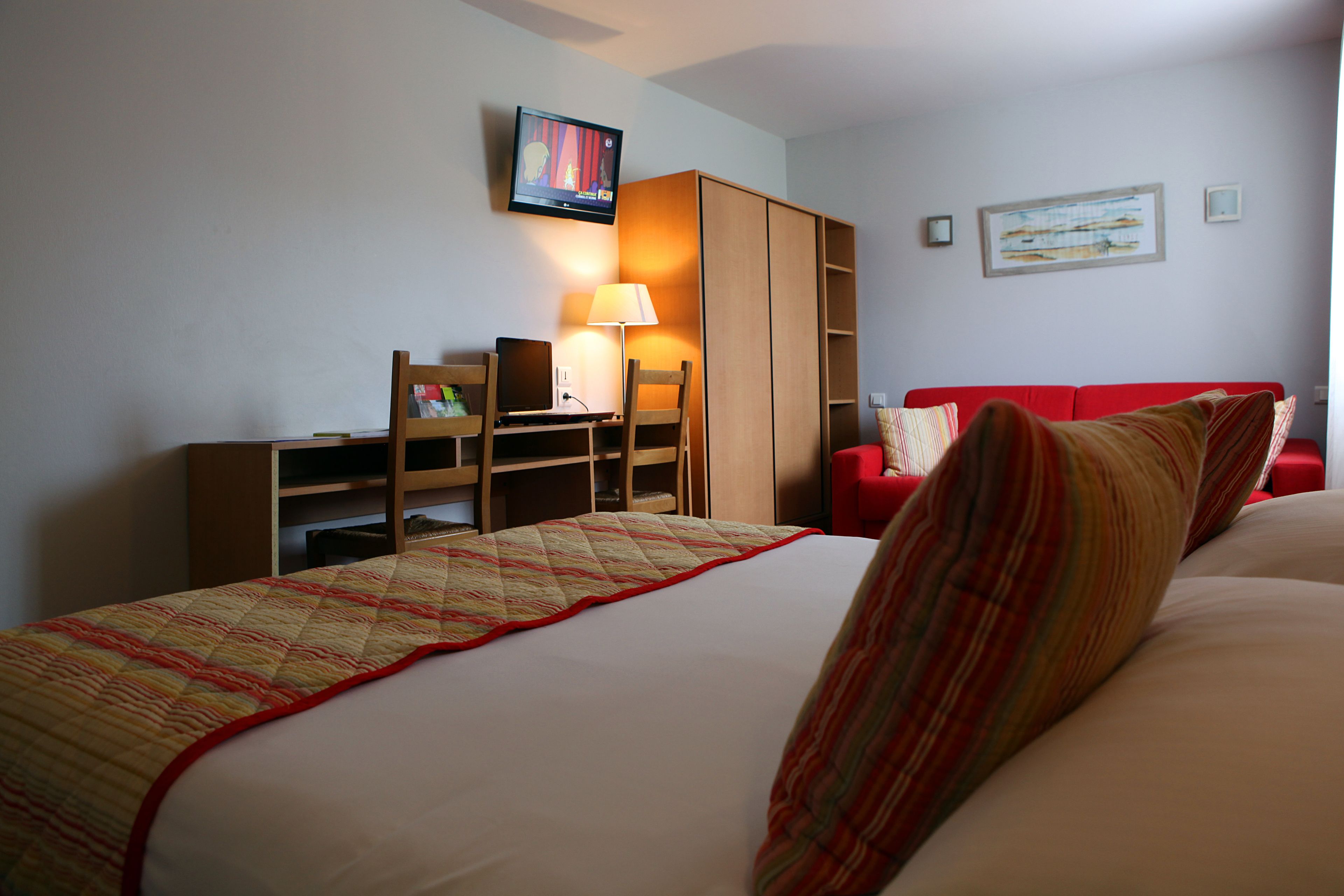 A hotel room with a bed, a desk with a computer, a TV, and a red sofa.